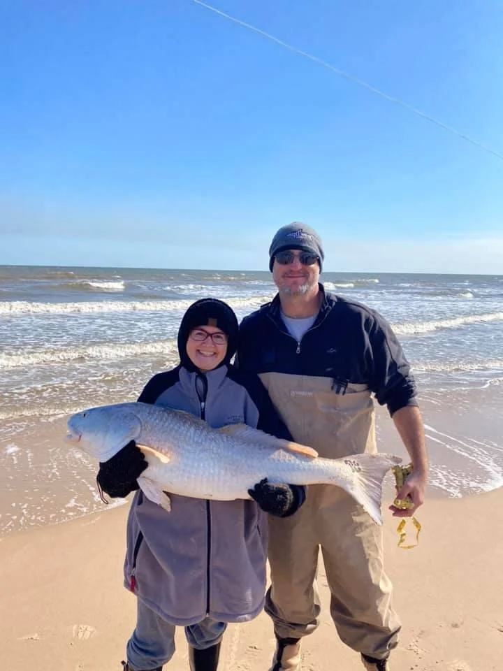 A smiling woman and man at the beach holding a large fish. The woman is wearing glasses, a gray jacket, and black gloves, and the man is wearing a beanie, sunglasses, and outdoor clothing. The ocean with small waves is in the background under a clear blue sky.