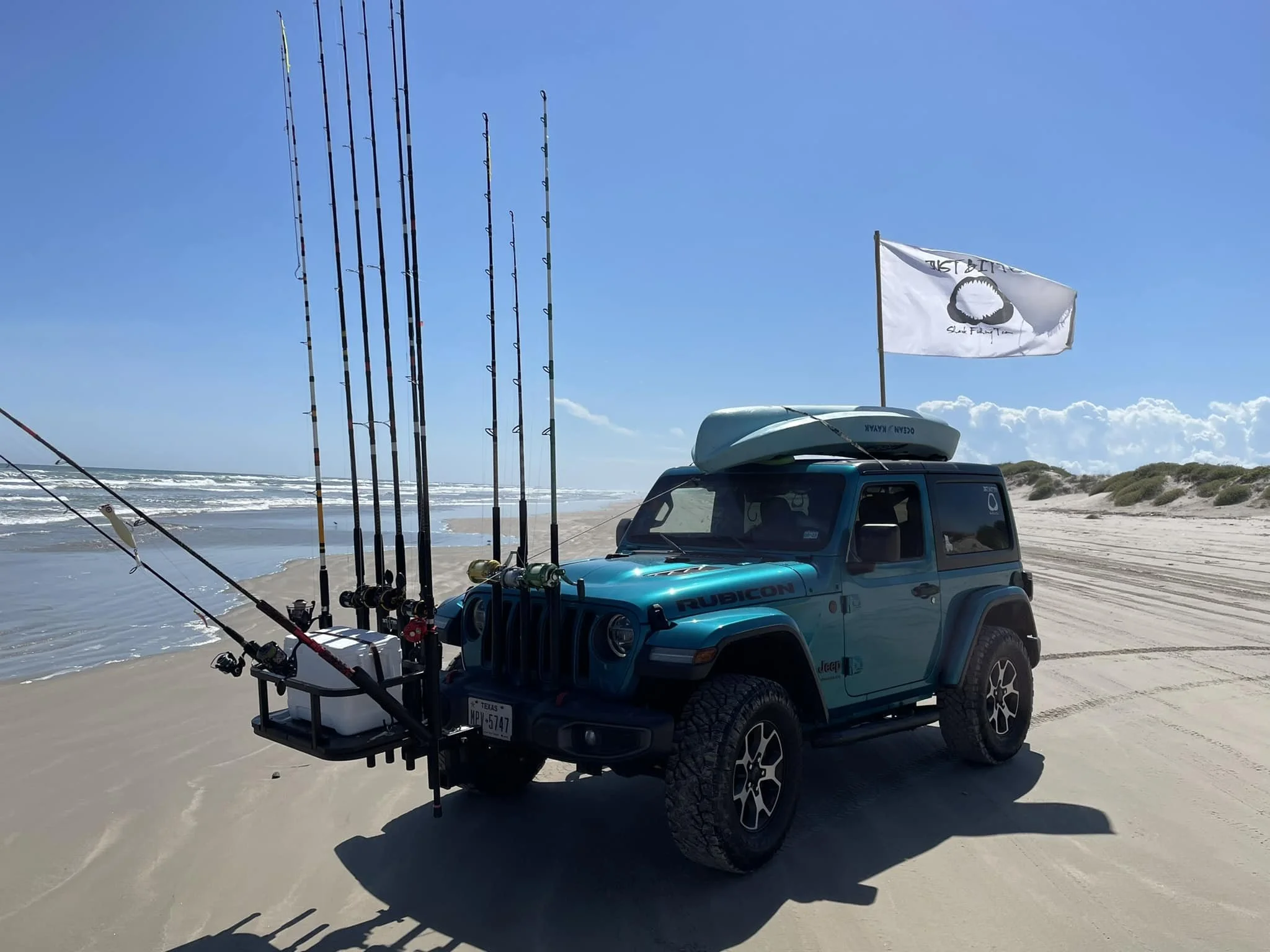 A blue Jeep Rubicon with a kayak and fishing rods attached, parked on a sandy beach with the ocean in the background under a clear blue sky. A white flag with a black circle and text is mounted on the vehicle.
