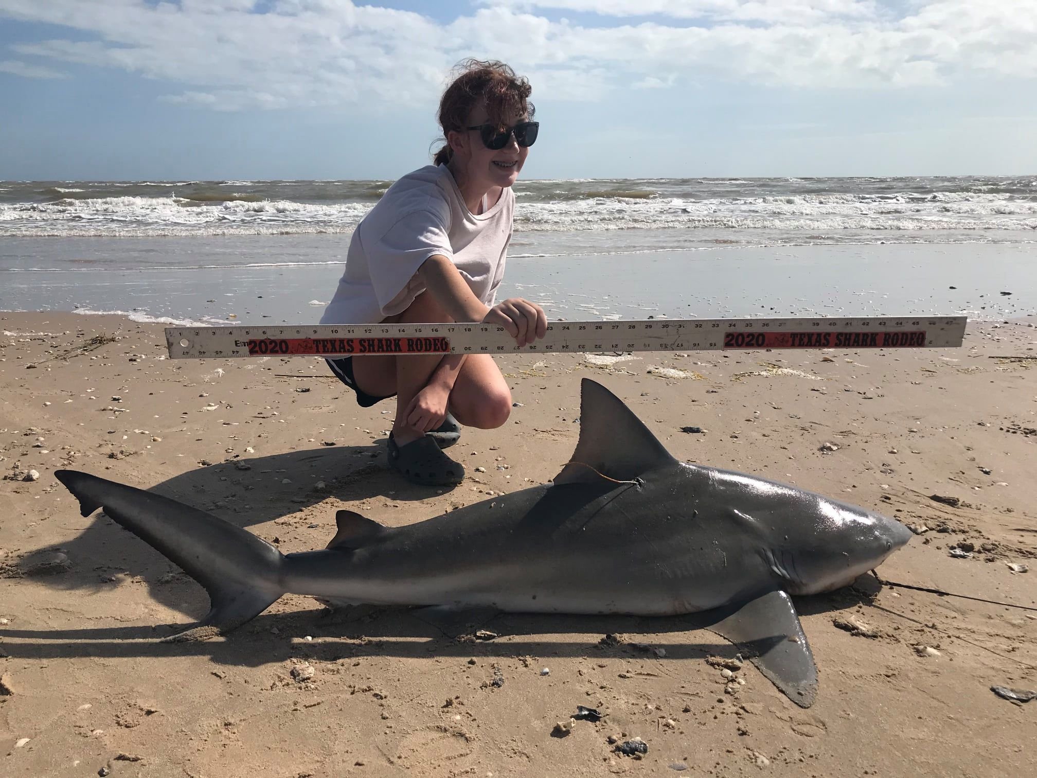 A woman with sunglasses kneels on a sandy beach next to a large dead shark, holding a measuring stick that reads "2020 Texas Shark Rodeo" with the ocean in the background.