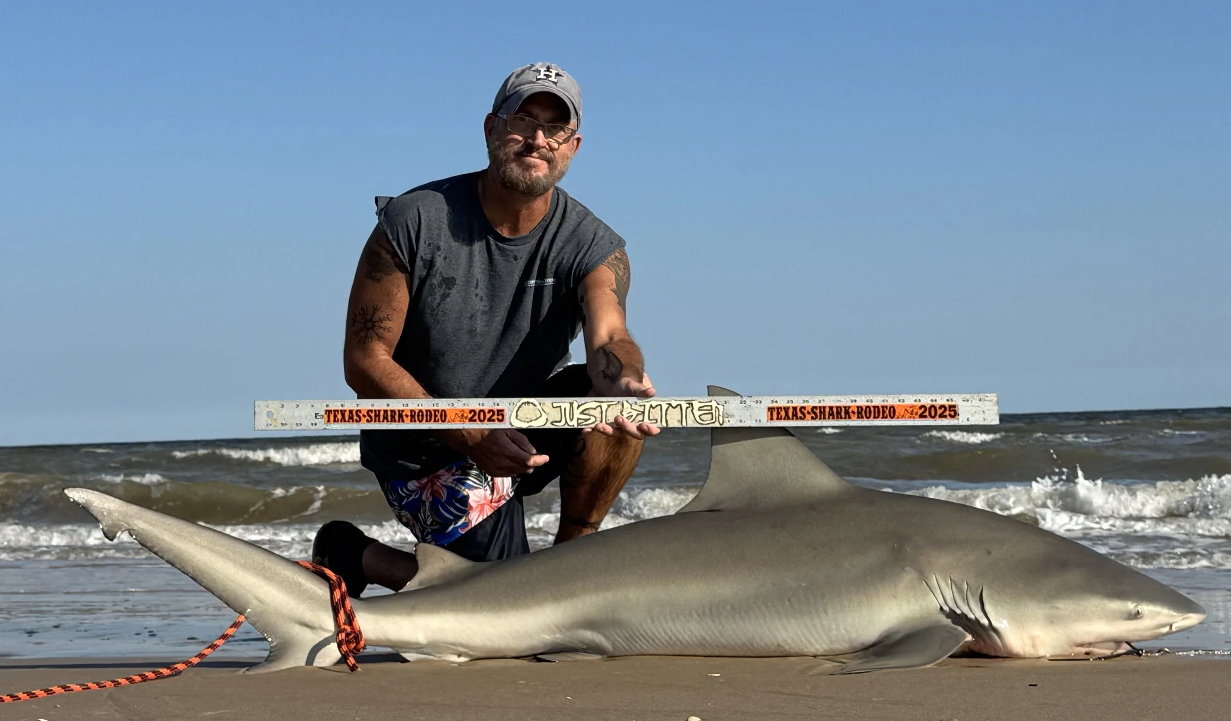 A man kneeling on the beach holding a measuring stick over a large shark lying on the sand with ocean waves in the background.