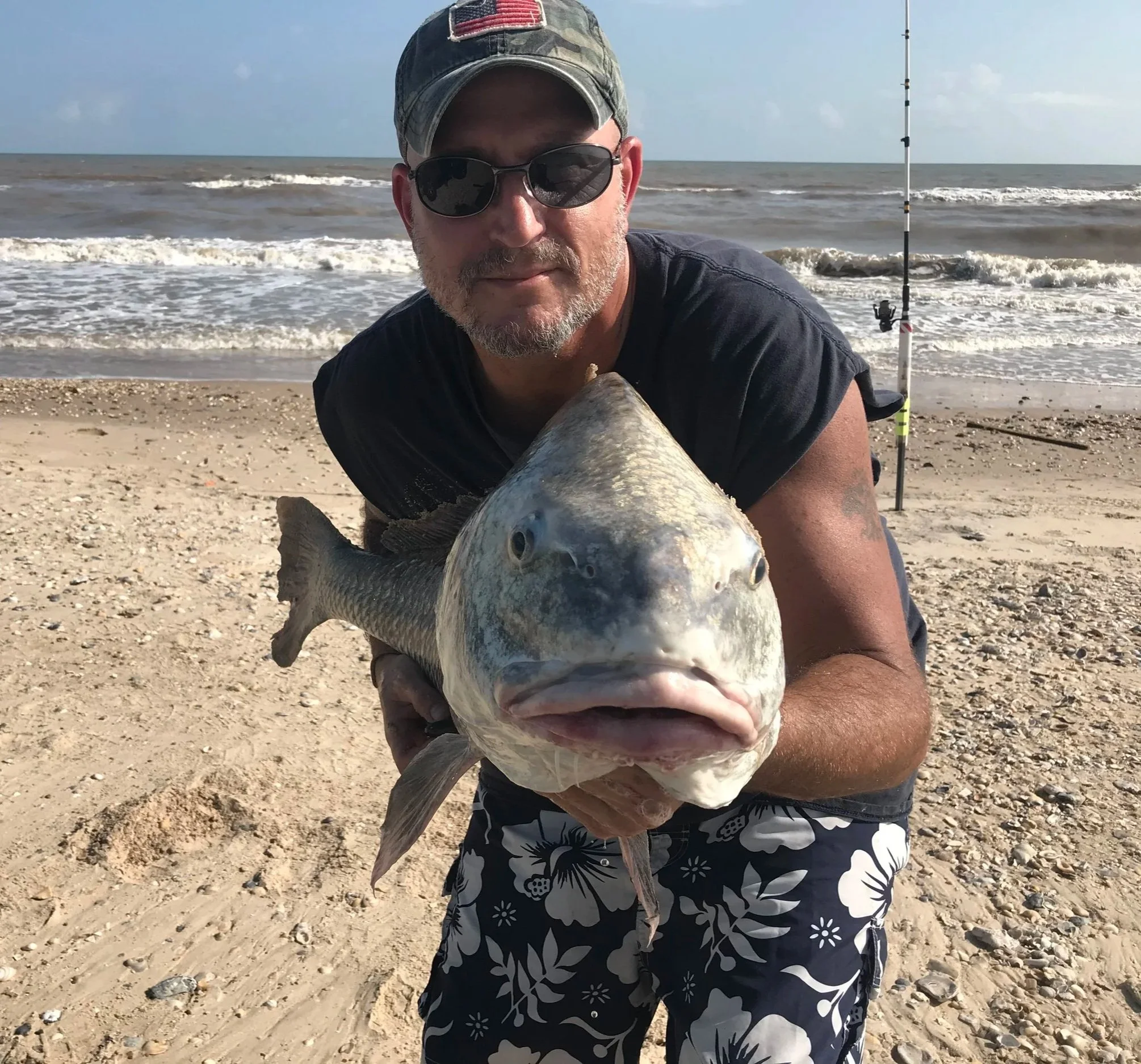 Man wearing sunglasses and a camouflage cap holding a large fish on a sandy beach with ocean waves in the background.