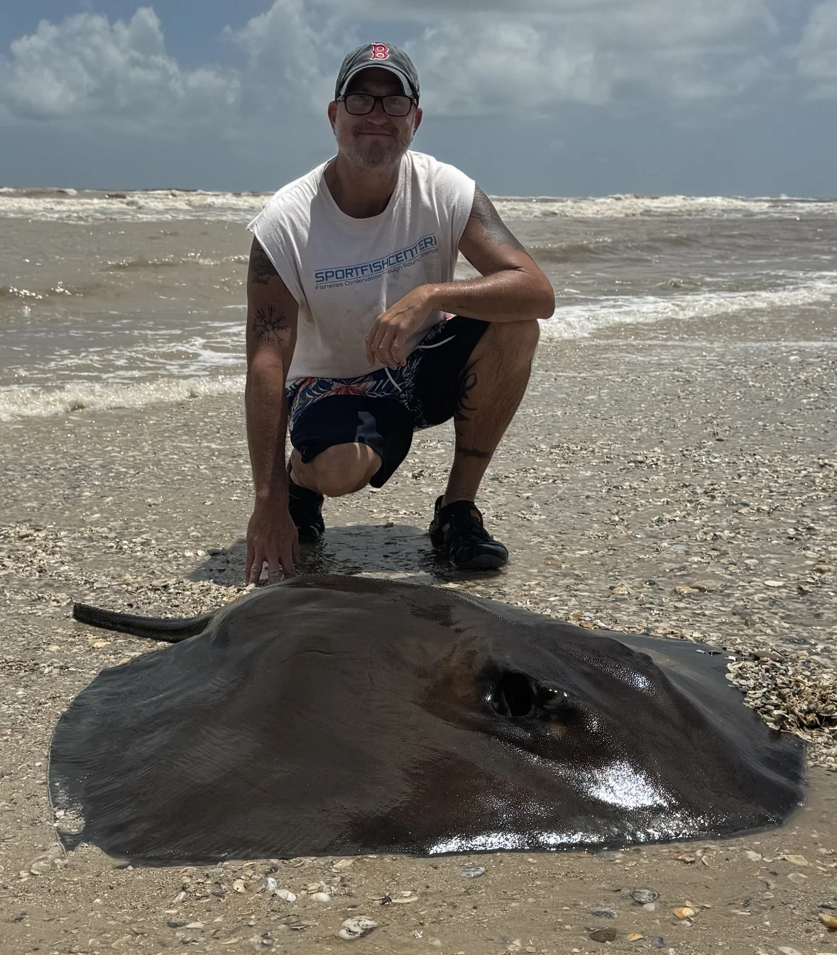 A man crouches on a beach next to a large stingray lying on the sand. The man is wearing a gray cap, glasses, a white sleeveless t-shirt, black shorts, and black shoes. The beach has shells and seashell fragments, and the ocean with waves can be seen in the background under a cloudy sky.