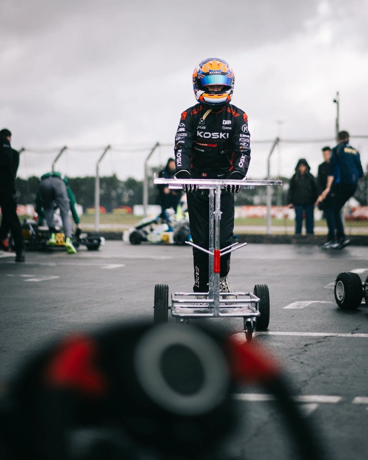 A race car driver in a racing suit and helmet standing on a tire cart at a racing track, with several other people and go-karts in the background during overcast weather.