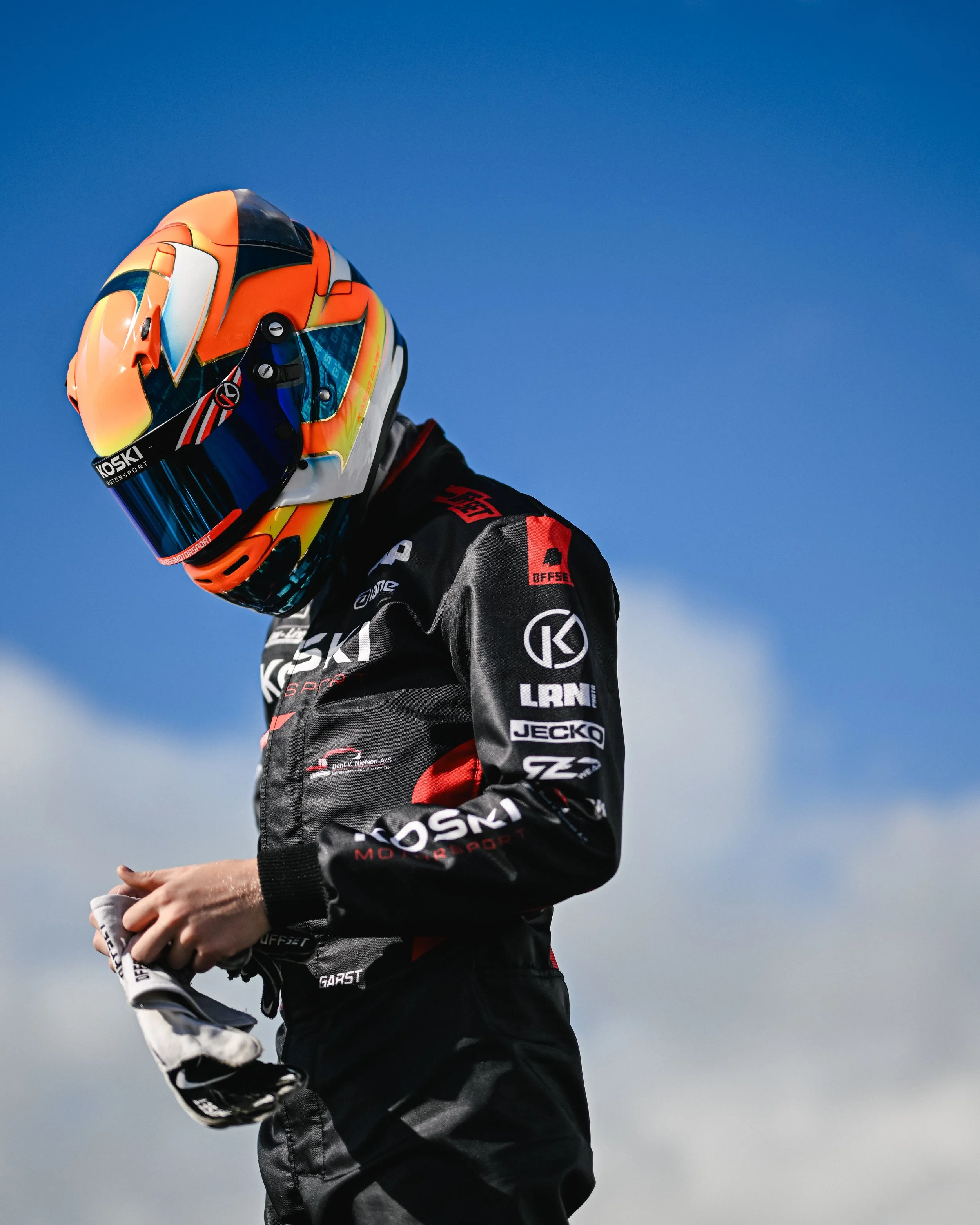 A race car driver in a black and red racing suit and a colorful helmet standing against a blue sky background.
