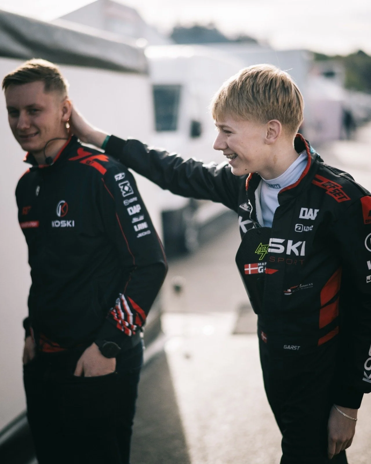 Two young men in racing suits standing near each other, one patting the other on the head, in a racing paddock area.