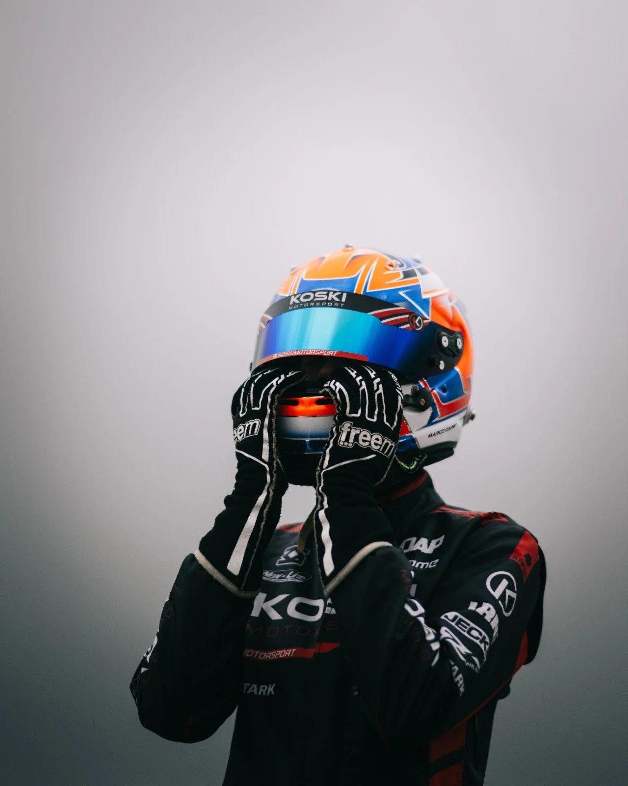 Race car driver in black racing suit and gloves, holding a colorful racing helmet against a plain gray background.