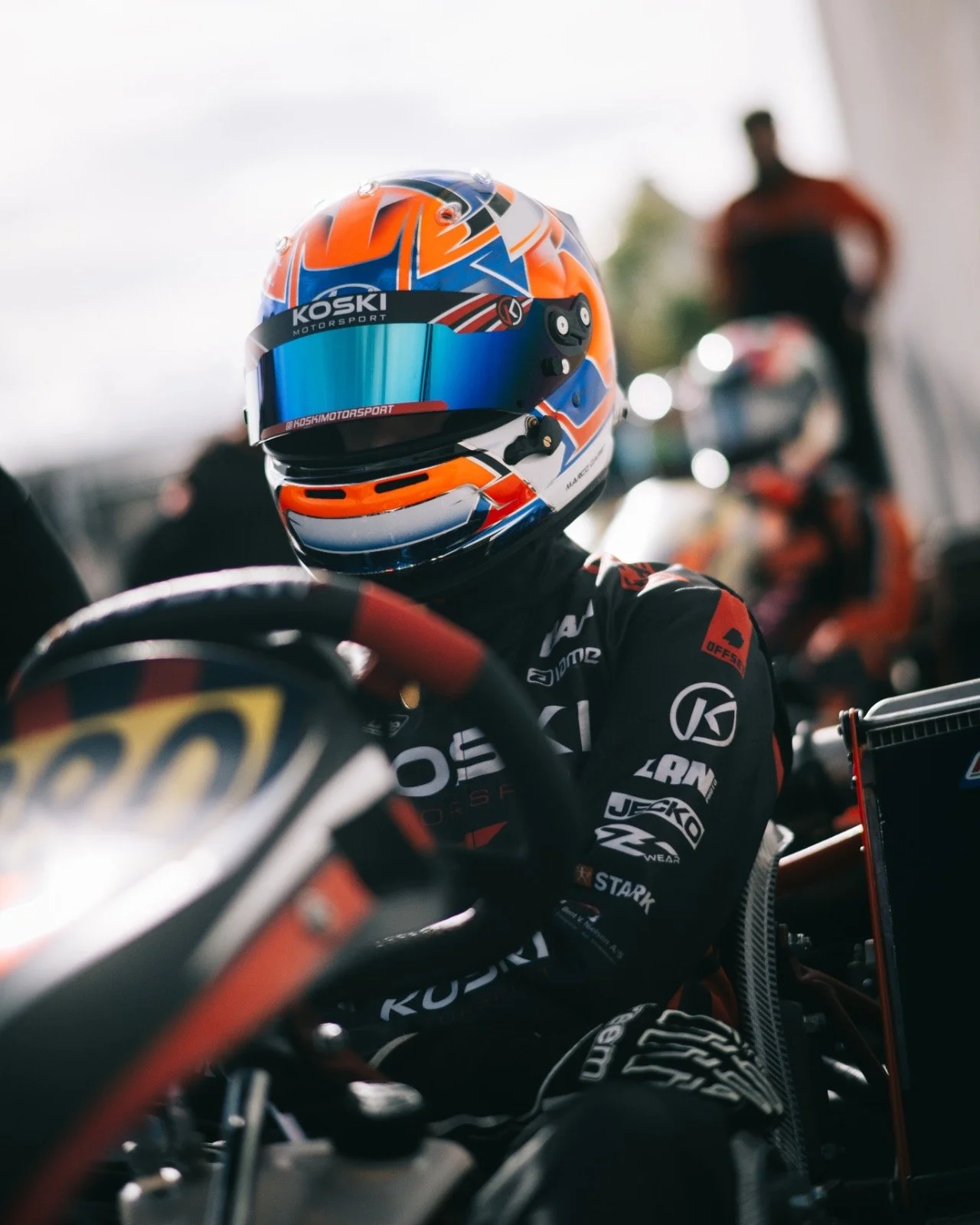 Close-up of a race car driver sitting in a race car, wearing a colorful helmet with a blue visor and racing suit with sponsor logos. The background shows a blurred person standing near a motorcycle.