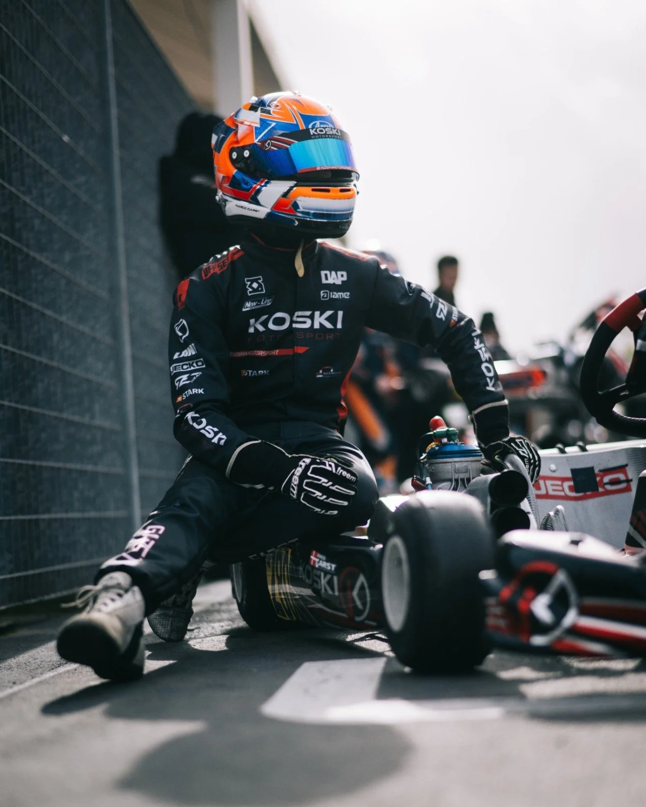A race car driver in a racing suit and helmet kneeling beside a formula racing car in a pit stop area.