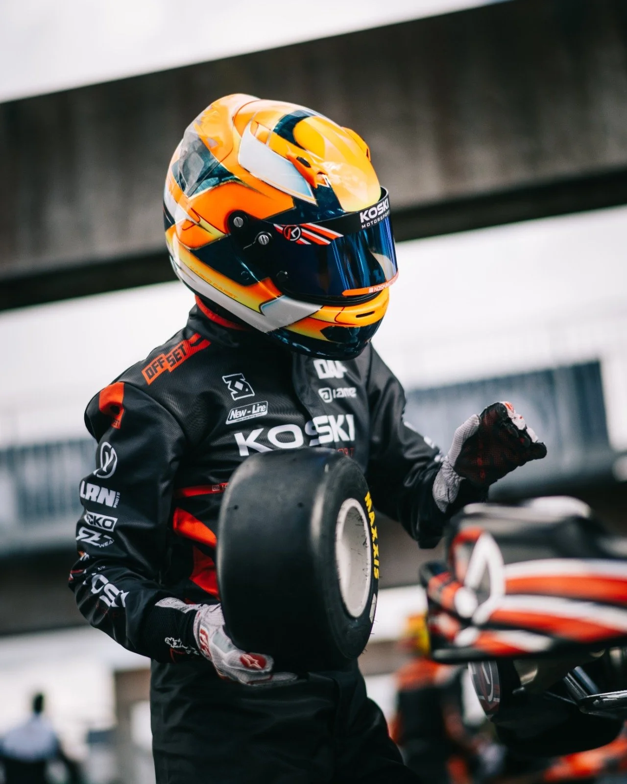 A race car driver in a black racing suit with various sponsor logos, holding a racing tire, wearing a colorful orange and yellow helmet, and standing near a racing motorcycle.
