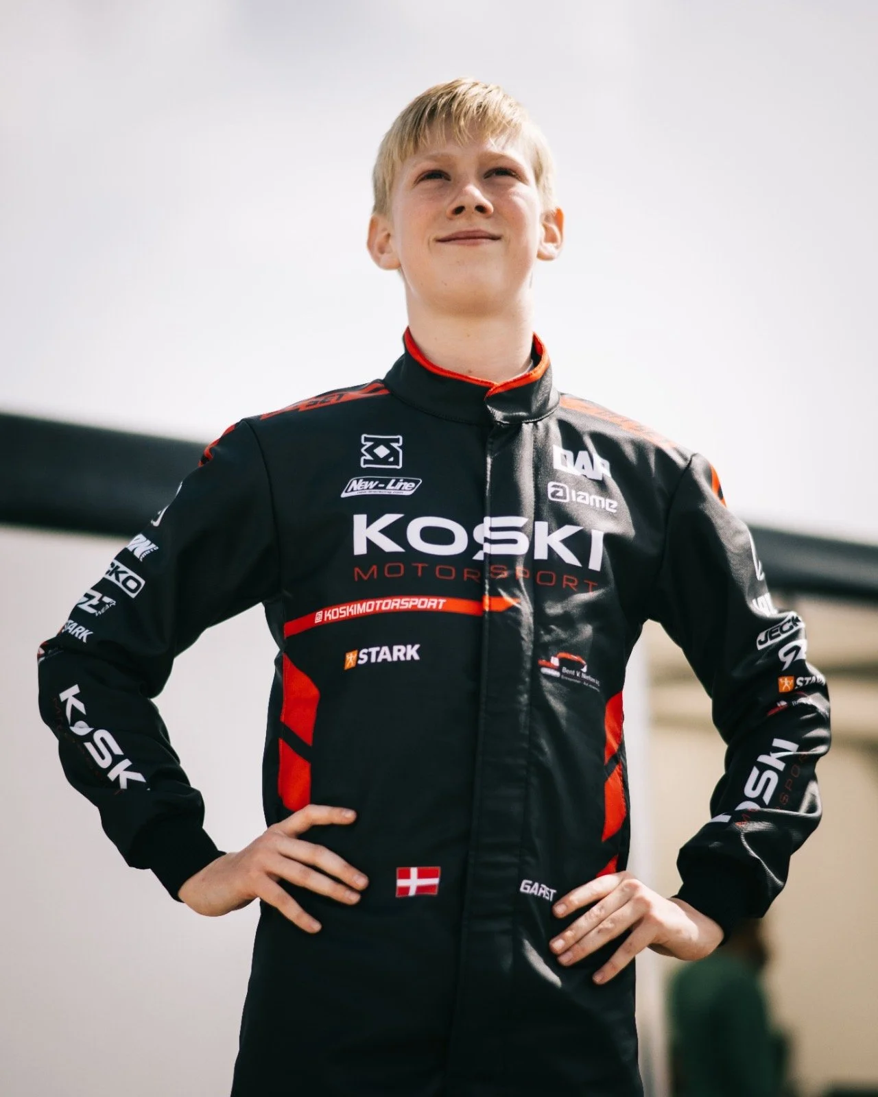 Young male race car driver in black racing suit with red accents, standing with hands on hips, looking confident outdoors.
