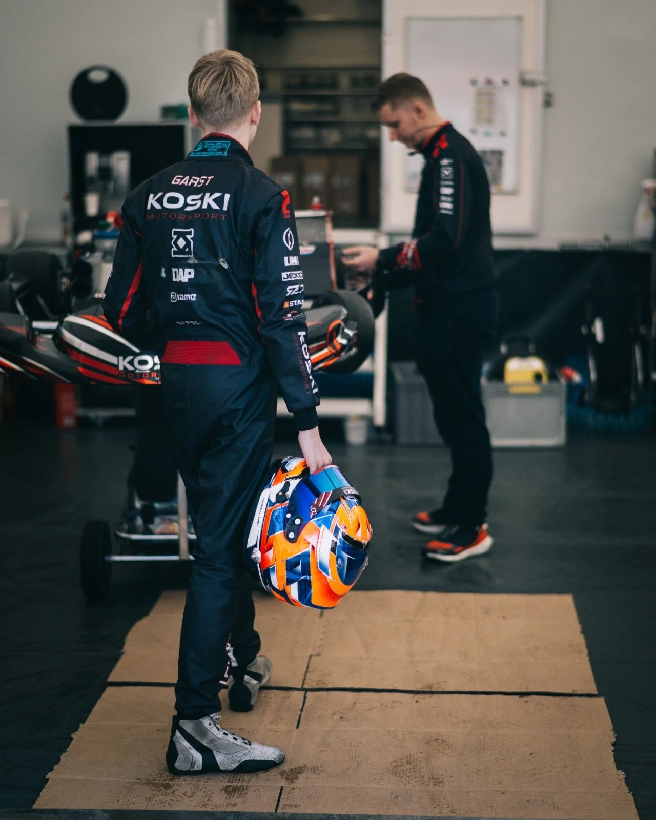 Two young race car drivers in racing suits preparing in a garage, one holding a colorful helmet and the other working on a race car in the background.