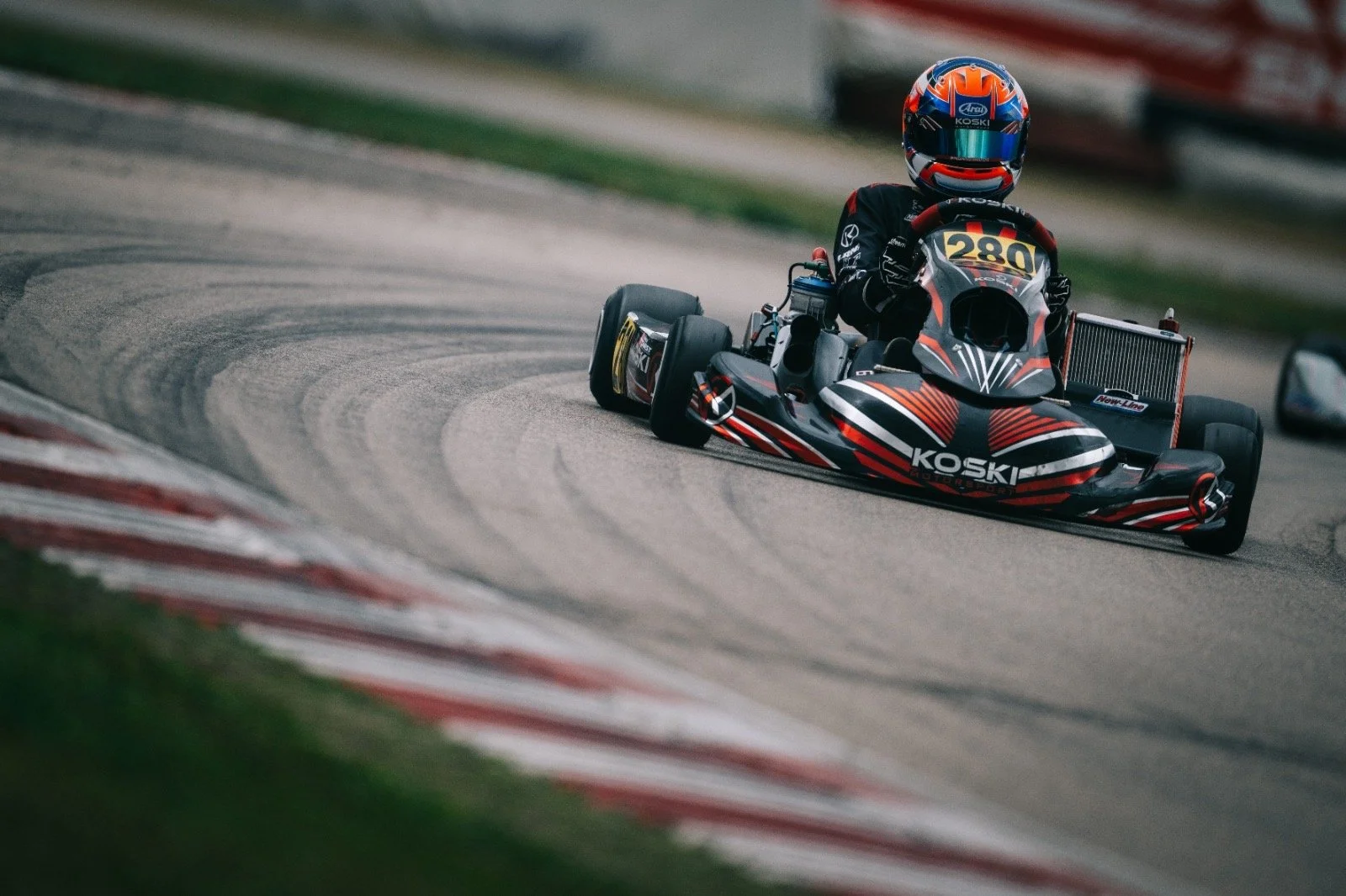 A person in racing gear driving a go-kart on a race track, wearing a blue and red helmet, with the go-kart labeled 'KOSKI' and number 280.