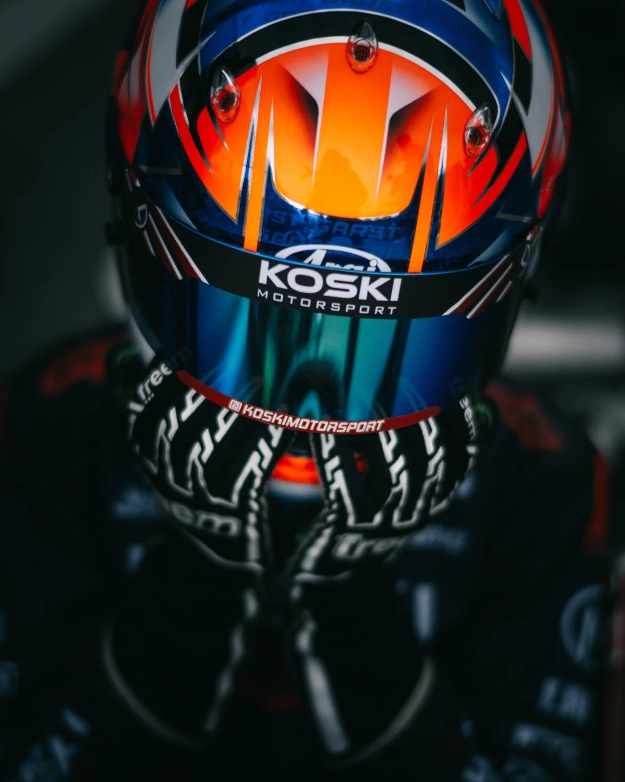 Close-up of a race car driver wearing a racing helmet with vibrant orange, blue, and black colors, and the text 'KOSKI MOTORSPORT' on it. The driver is holding their gloves around their chin.