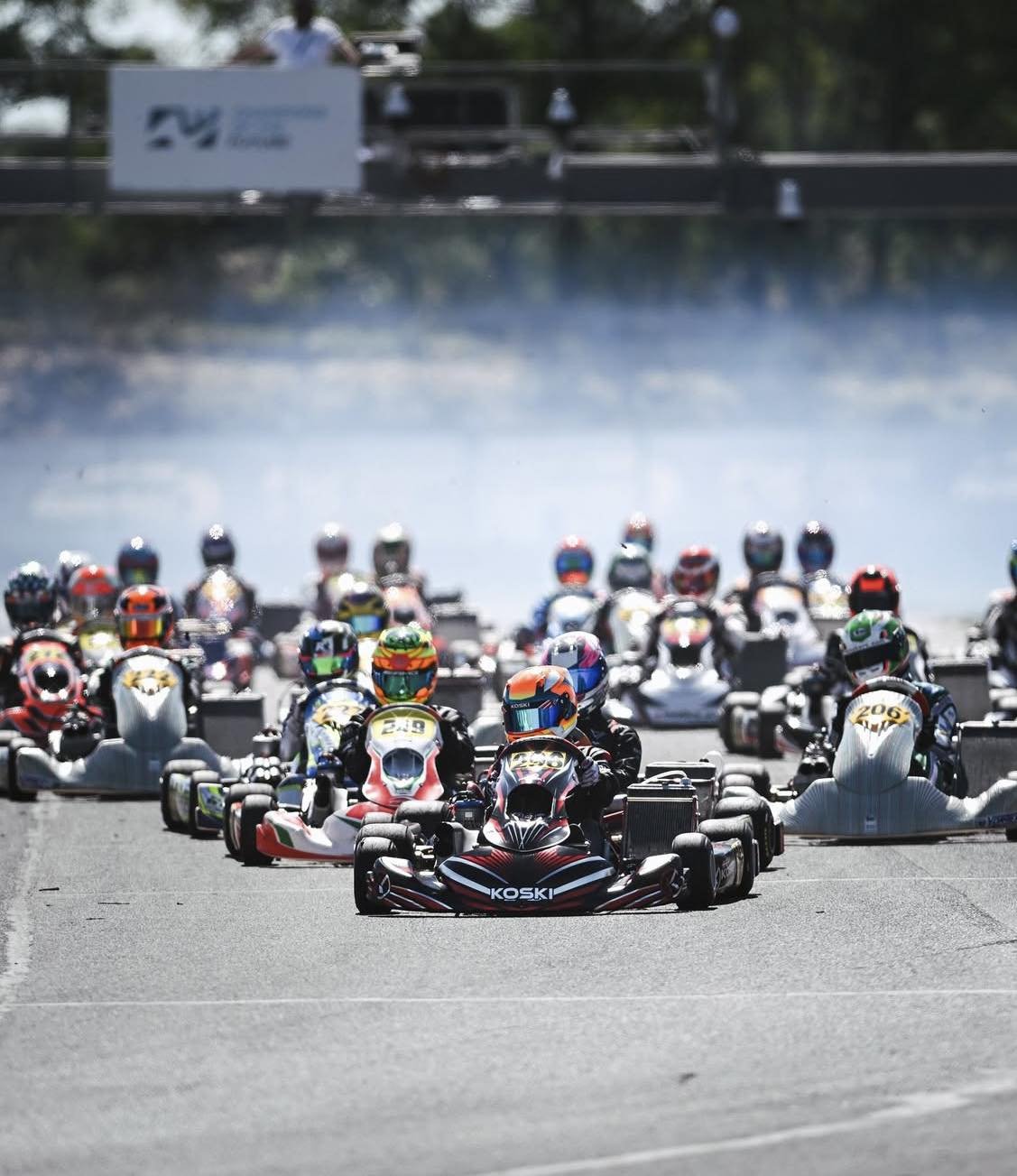 Go-kart race with numerous racers wearing colorful helmets and suits, racing on an outdoor track with a blurred background.