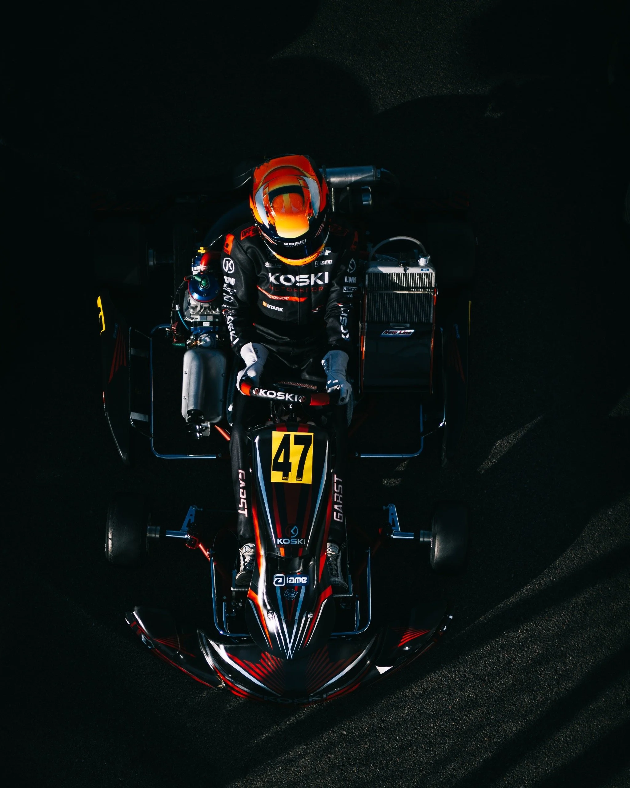 Top-down view of a race car driver sitting in a go-kart on a dark track, wearing a black racing suit and orange helmet with a reflective visor, with the number 47 displayed on the front of the kart.