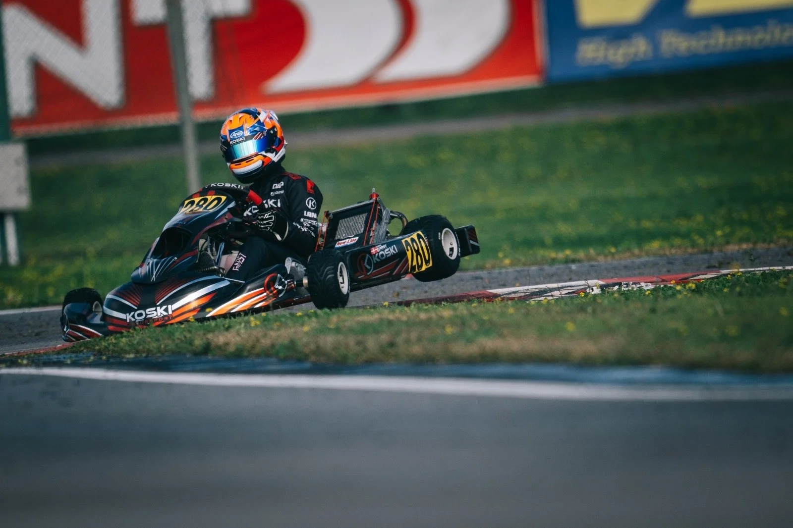 A person racing in a go-kart on a track, wearing a helmet and racing suit, with a blurred background of advertising banners and a grassy area.