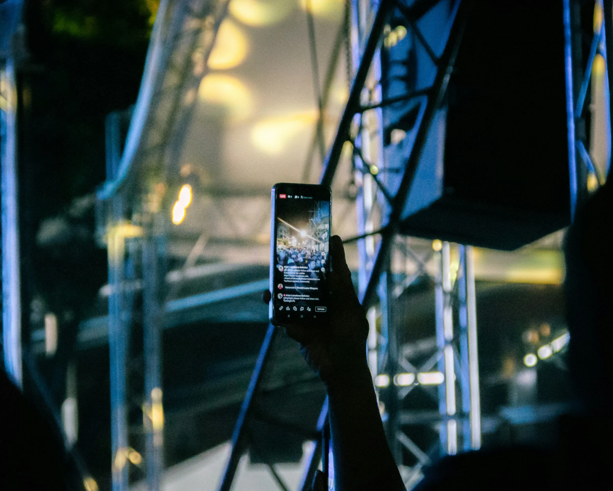 Person holding a smartphone recording a concert stage with scaffolding and lighting at night.