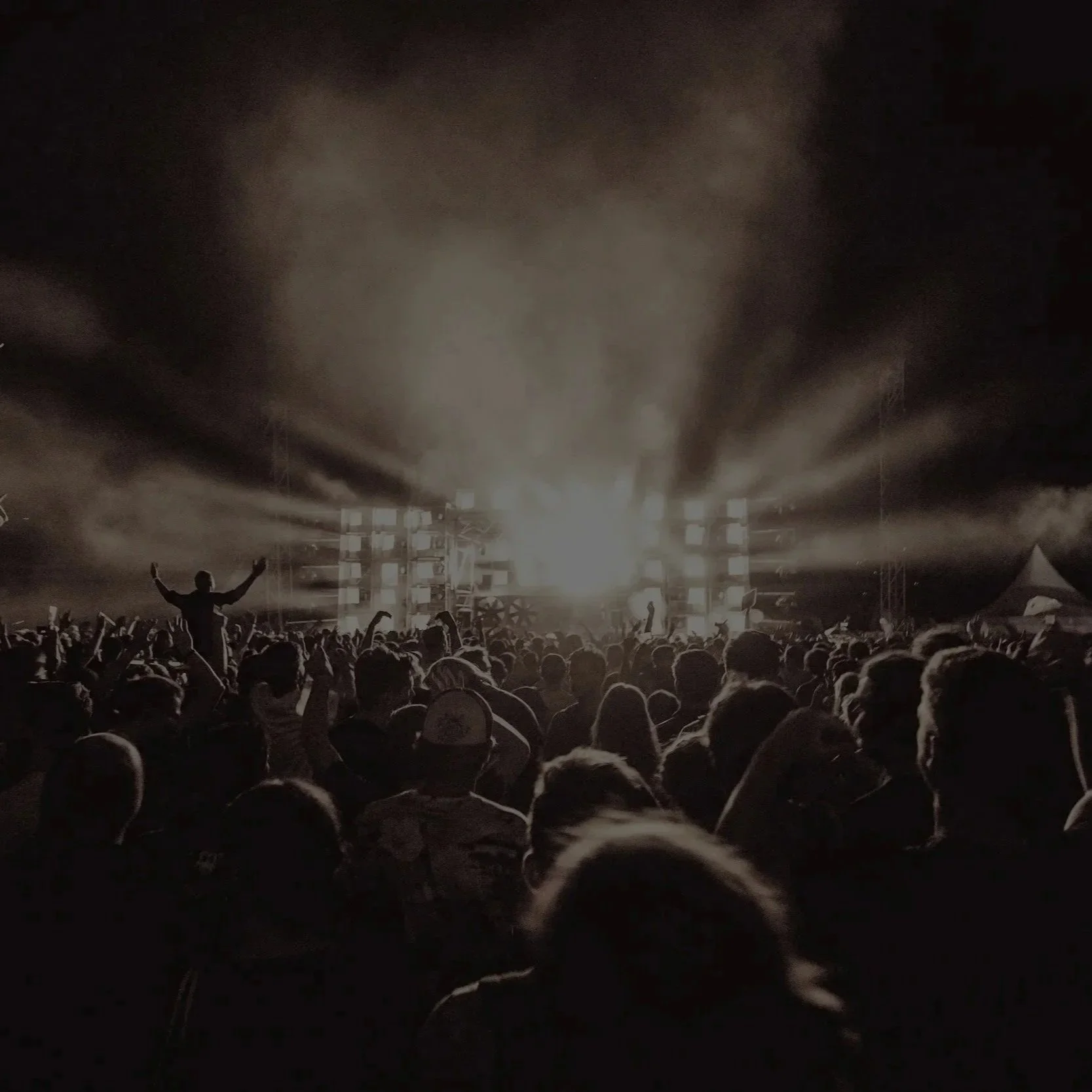A large crowd at a concert or music festival at night, with a stage illuminated by bright lights and smoke, and people dancing and having fun.