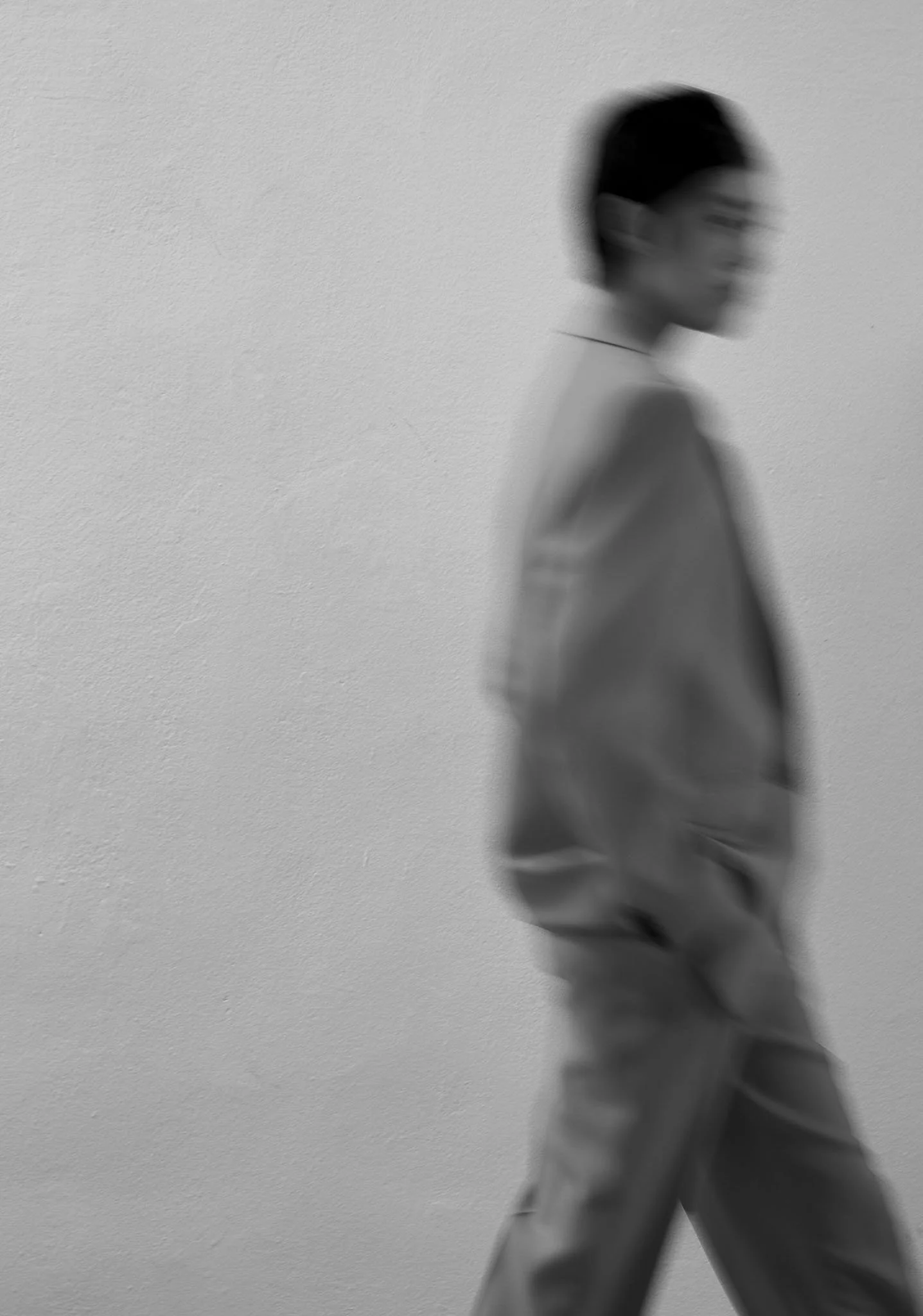 Blurry black and white photo of a person walking, facing right, with hands in pockets, against a plain wall background.