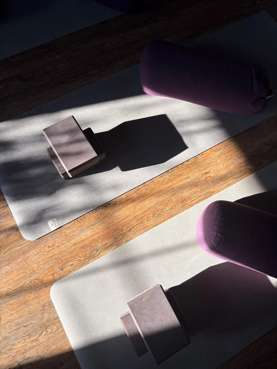Two yoga mats on a wooden floor with sunlight casting shadows, each with a foam block and purple bolster pillow.