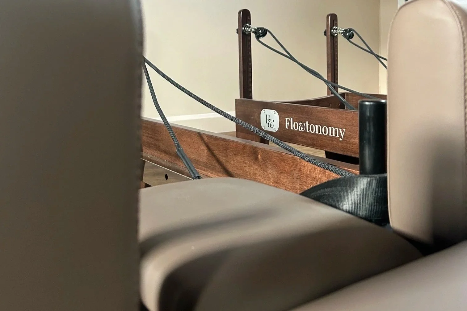 A close-up view of a wooden sign with the word 'Florotonomy' on it, seen through the gap between two beige chairs, with some black straps or cords attached to the sign.