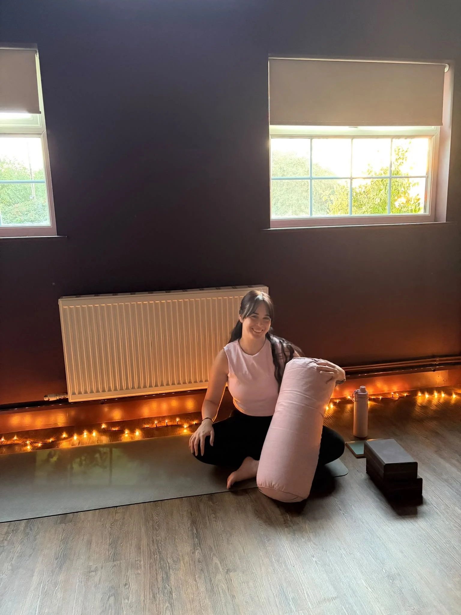 A woman sitting on the floor, smiling, holding a pink bolster, with a dark wall, two windows, and fairy lights along the wall in the background.