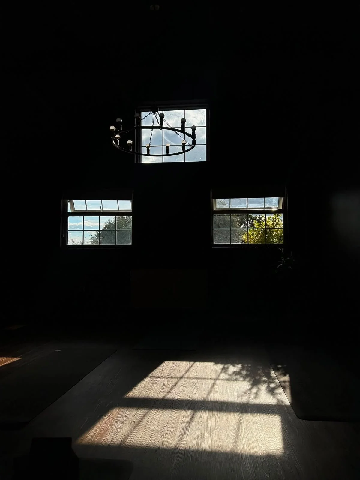 Dark room with sunlight coming through three windows, chandelier hanging from ceiling, and shadows cast on wooden floor.