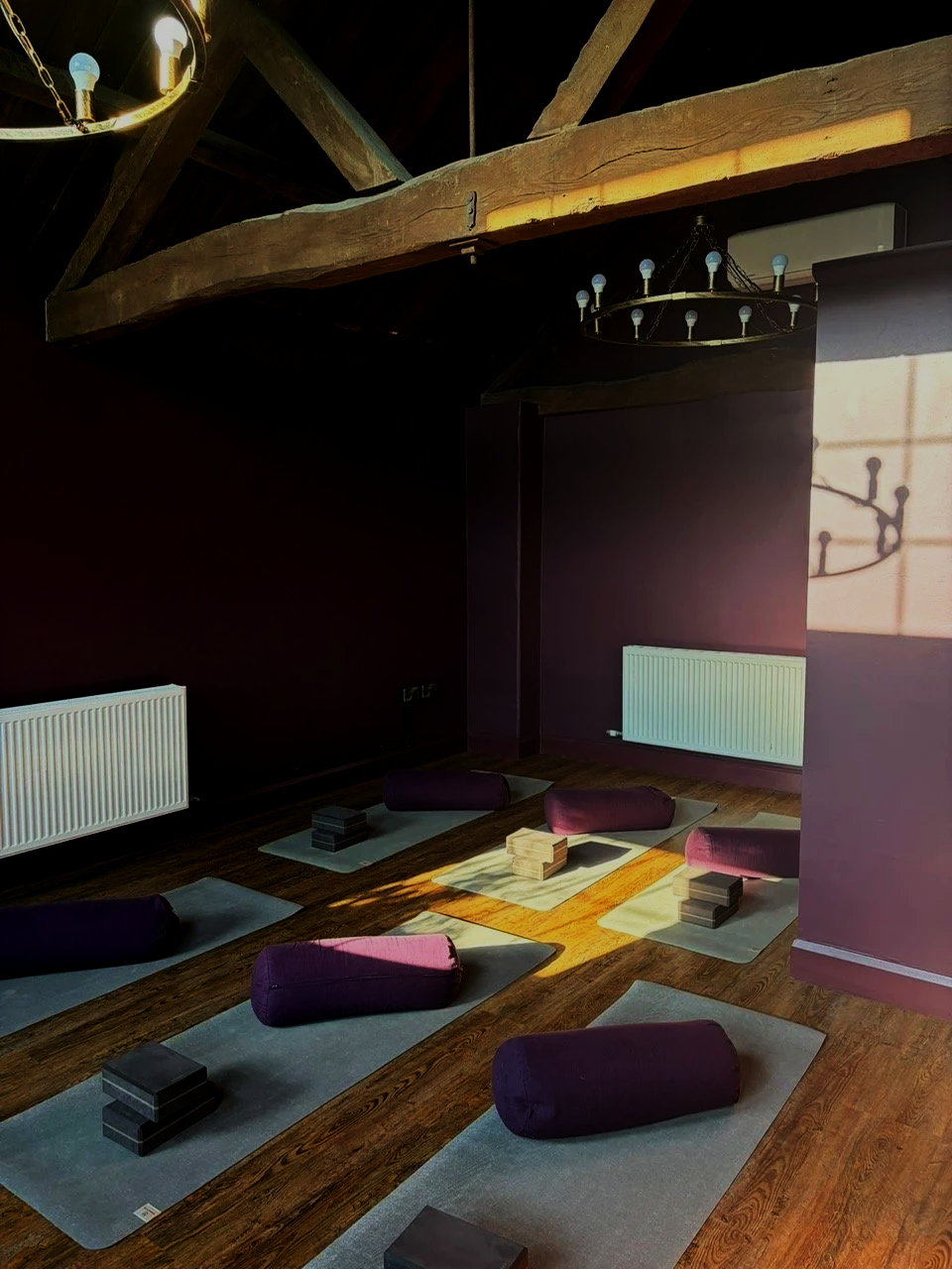 Yoga class setup with mats, bolsters, and blocks in a dimly lit room with wooden beams and chandeliers.