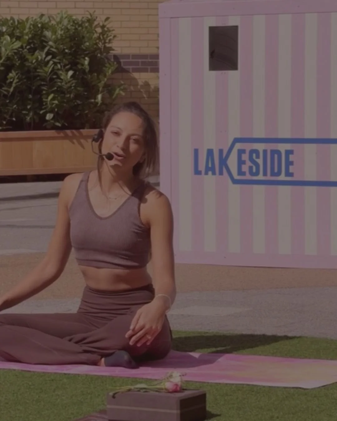 A woman sitting cross-legged on a yoga mat outdoors, wearing a microphone headset, with a pink and white striped background that reads 'LAKESIDE' and a brick wall with greenery behind her.