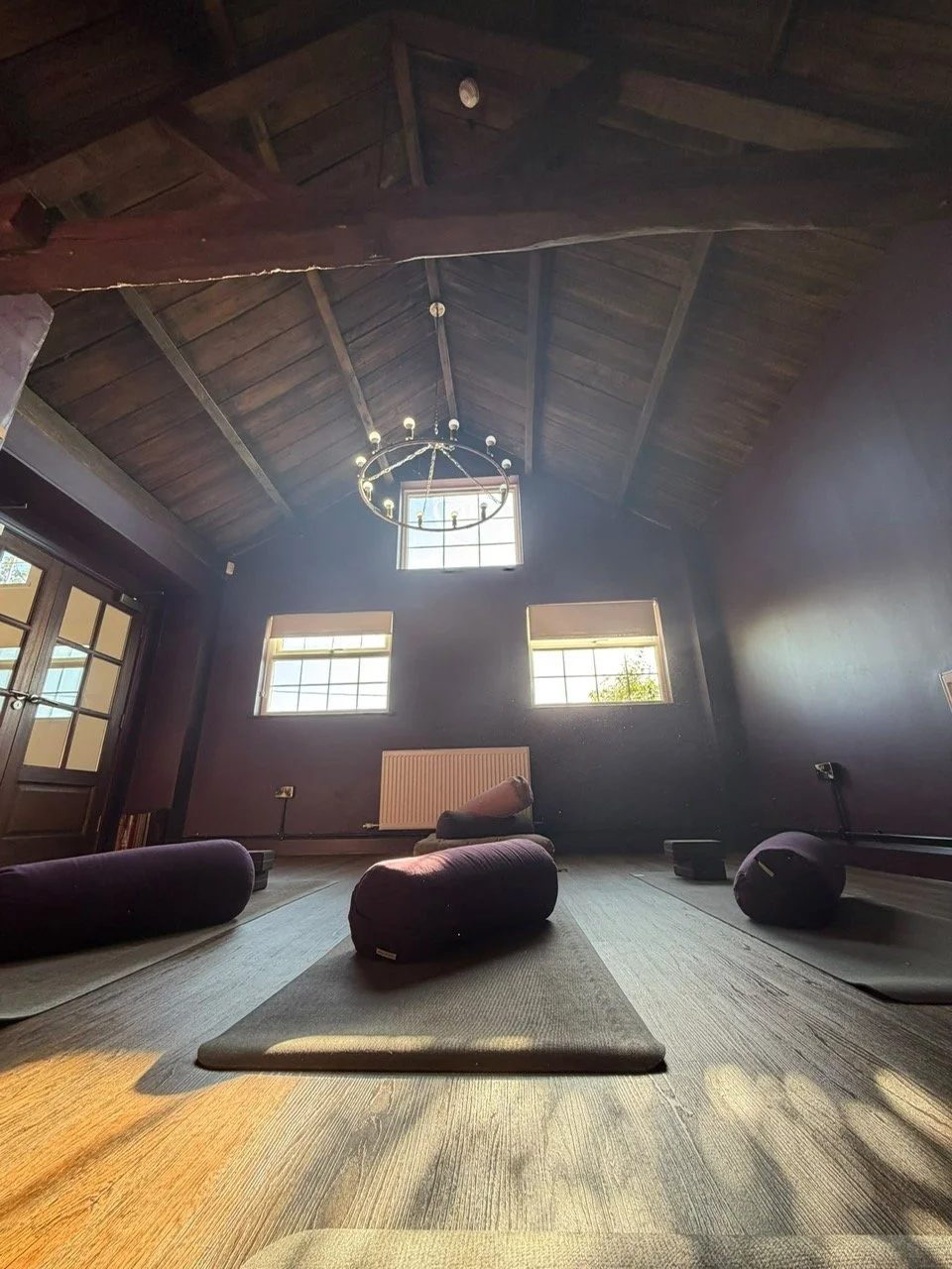 A yoga or meditation room with mats and bolsters, large windows, and a wooden ceiling with a chandelier.