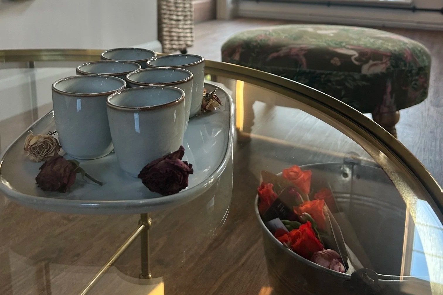 A glass-top table with six white cups and dried roses, next to a glass container filled with red roses, in a cozy indoor setting.
