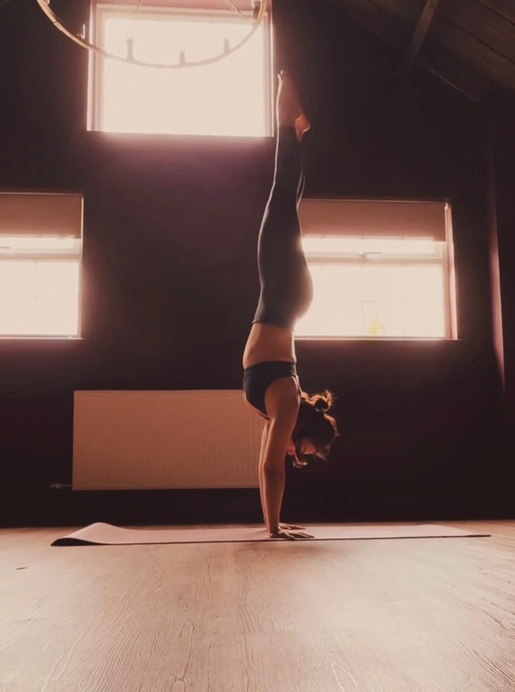 A woman practicing yoga indoors, performing a handstand on a yoga mat in a room with wooden floors and large windows allowing natural light.