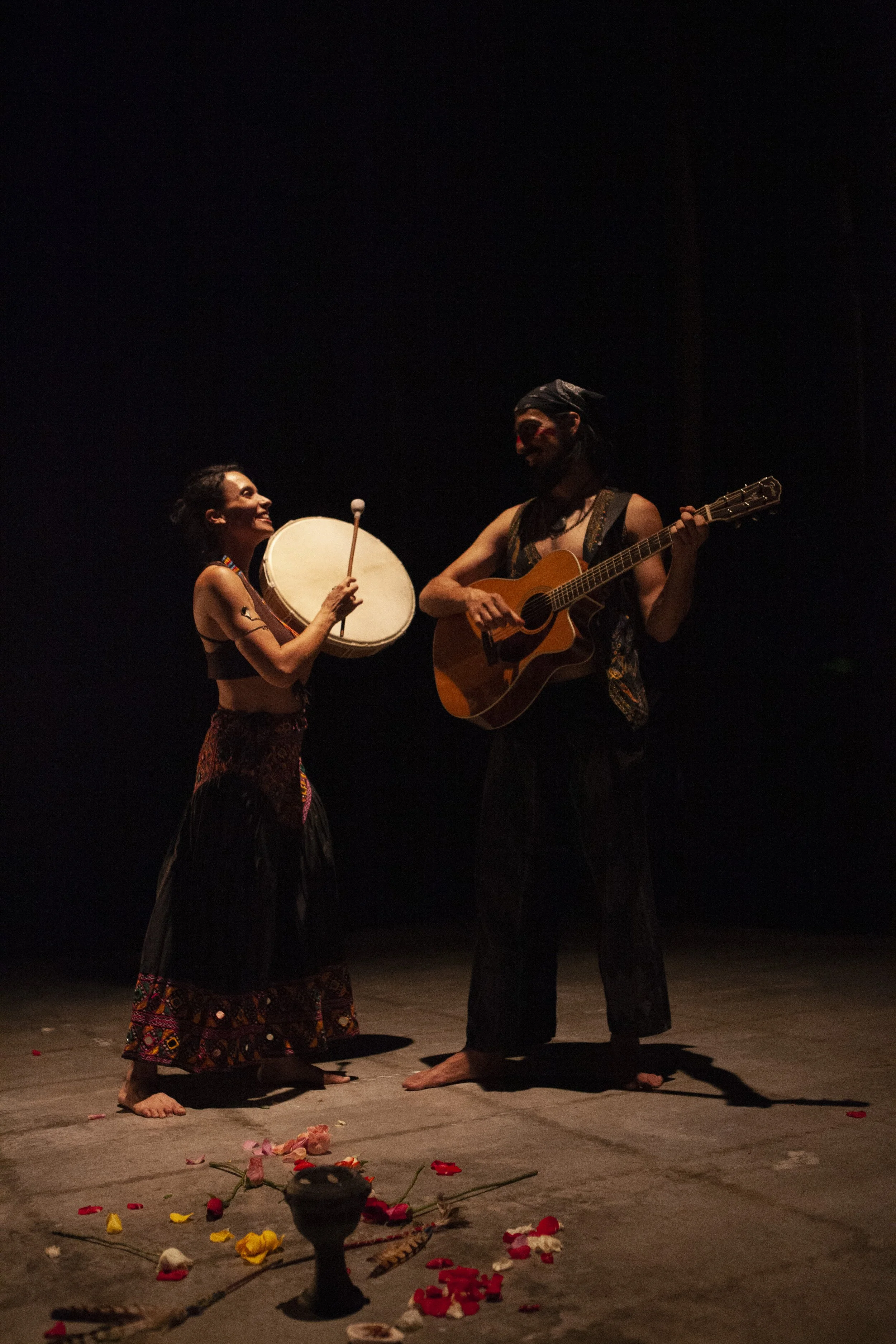 Two performers standing barefoot on a dimly lit stage, one playing guitar and the other holding a drumstick near a large drum, with flower petals scattered on the floor and a dark background.