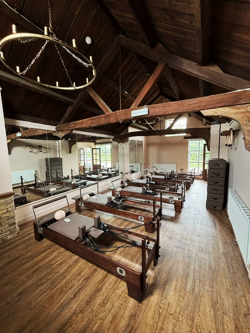 A Pilates studio with multiple Pilates reformer machines arranged in rows on wooden floors, with mirrors on the walls and a high wooden ceiling with exposed beams.