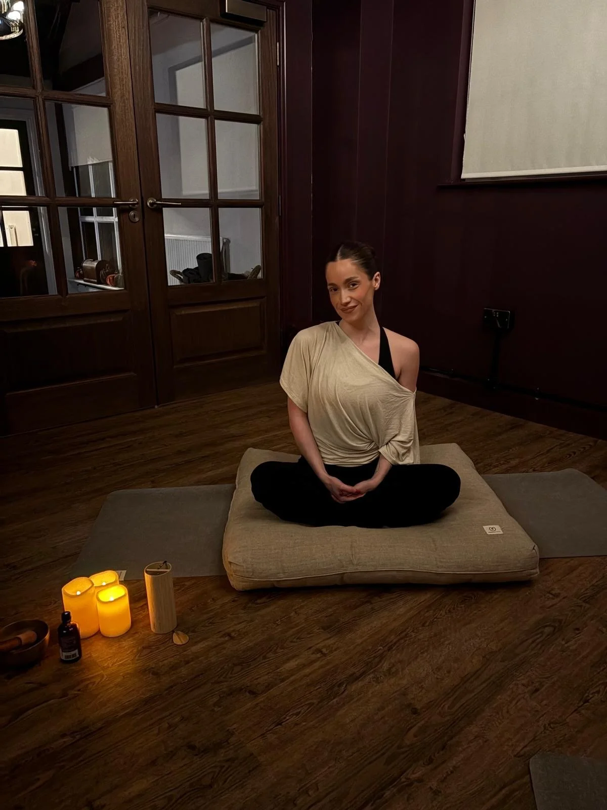 A woman sitting cross-legged on a beige cushion during a yoga or meditation session in a dimly lit room, with candles, essential oils, and candles on the floor nearby.