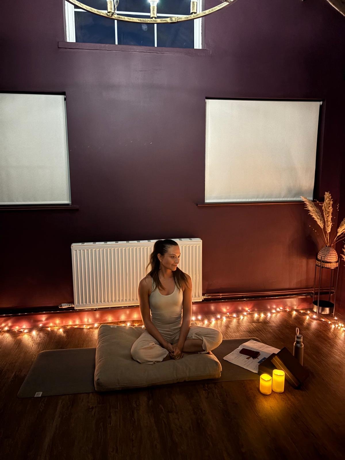 A woman is sitting cross-legged on floor cushion in a dimly lit room decorated with string lights and candles, with a dark maroon wall and two windows with closed blinds behind her.