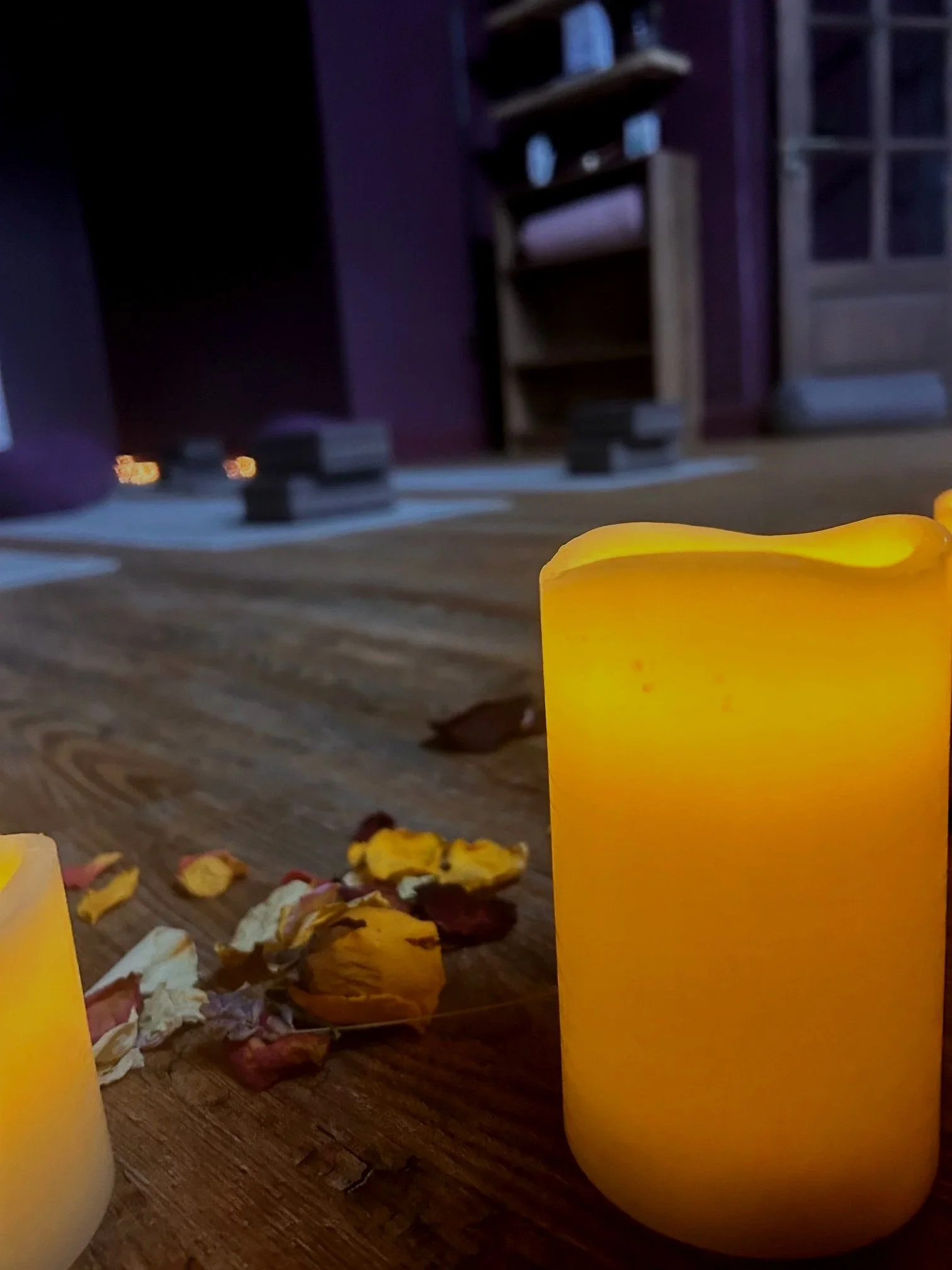 Yoga mats surrounded by candles and dried flowers, in a dimly lit room with purple walls and shelving in the background.