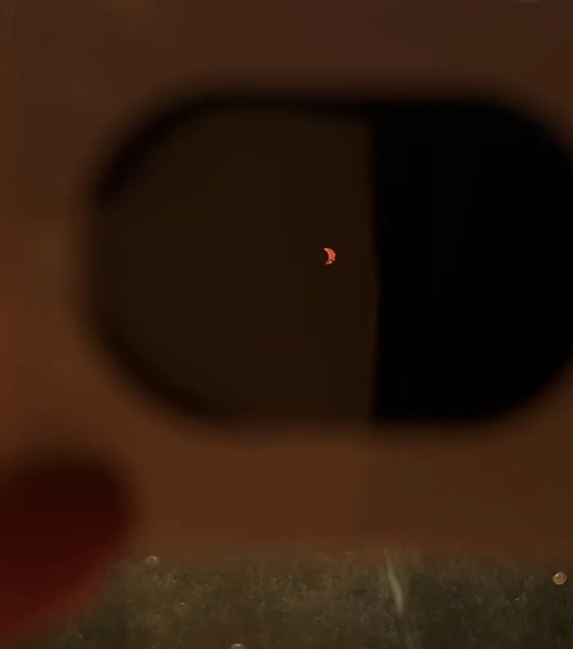 Close-up of a small, reddish orange crescent moon seen through a camera or telescope with a black background.