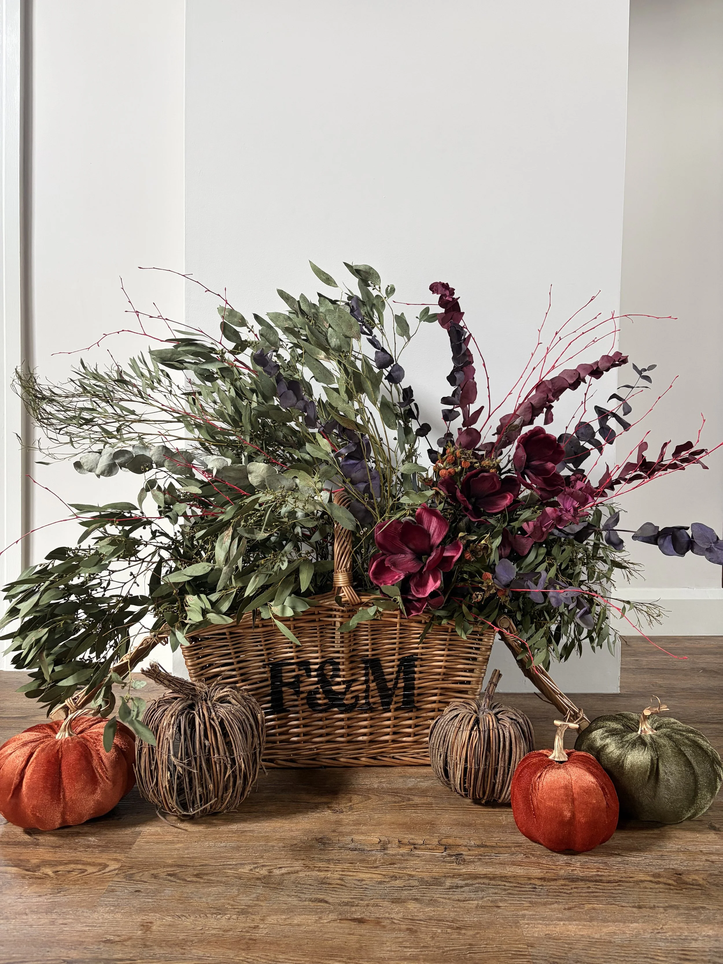 A decorative fall arrangement in a wicker basket with fall pumpkins on a wooden table inside a room.