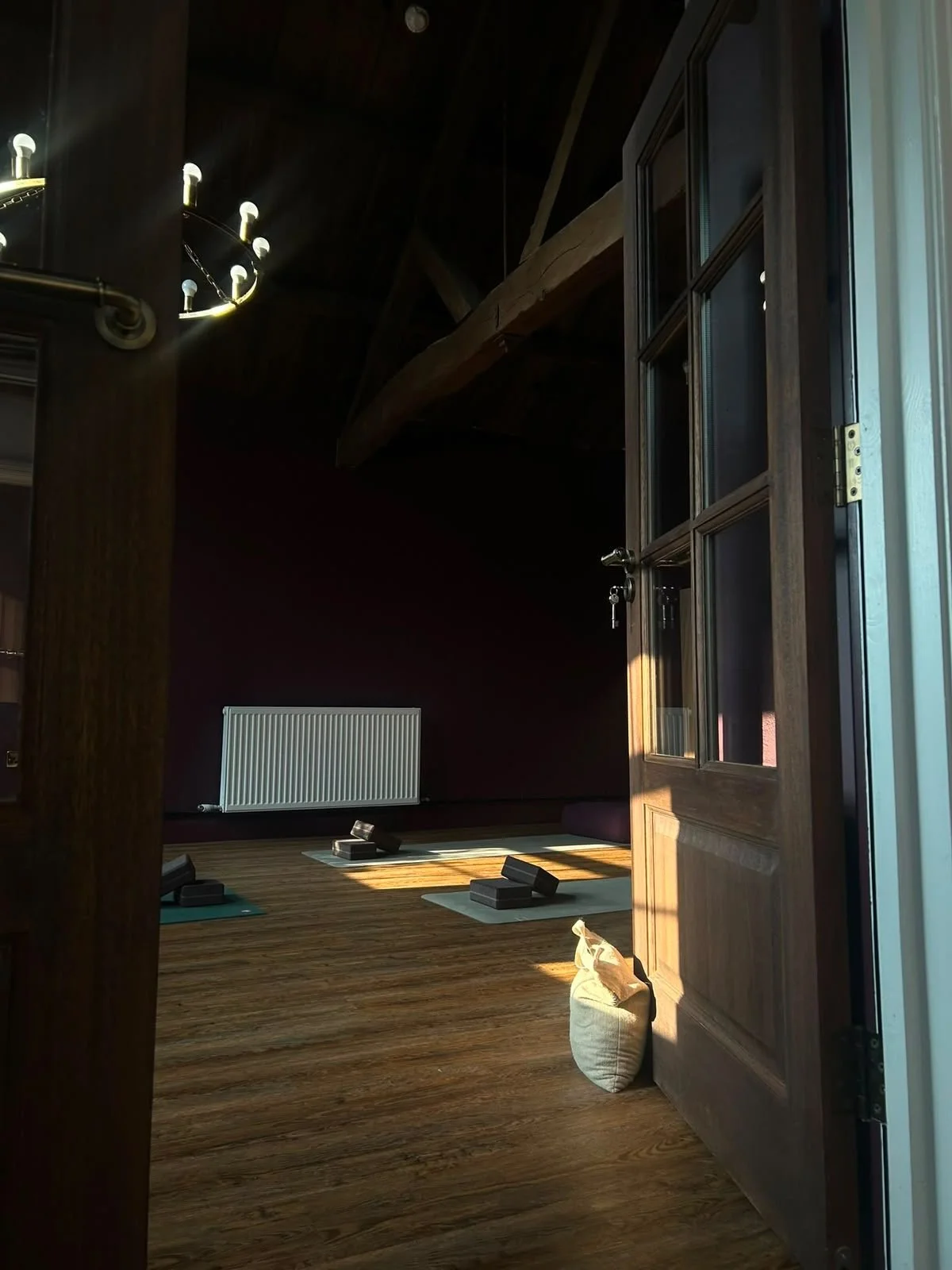 A cozy room set up for yoga or meditation with mats, blocks, and a bag, illuminated by natural sunlight near a wooden door with a glass window, wooden floor, a radiator on a dark burgundy wall, and a chandelier with exposed bulbs.