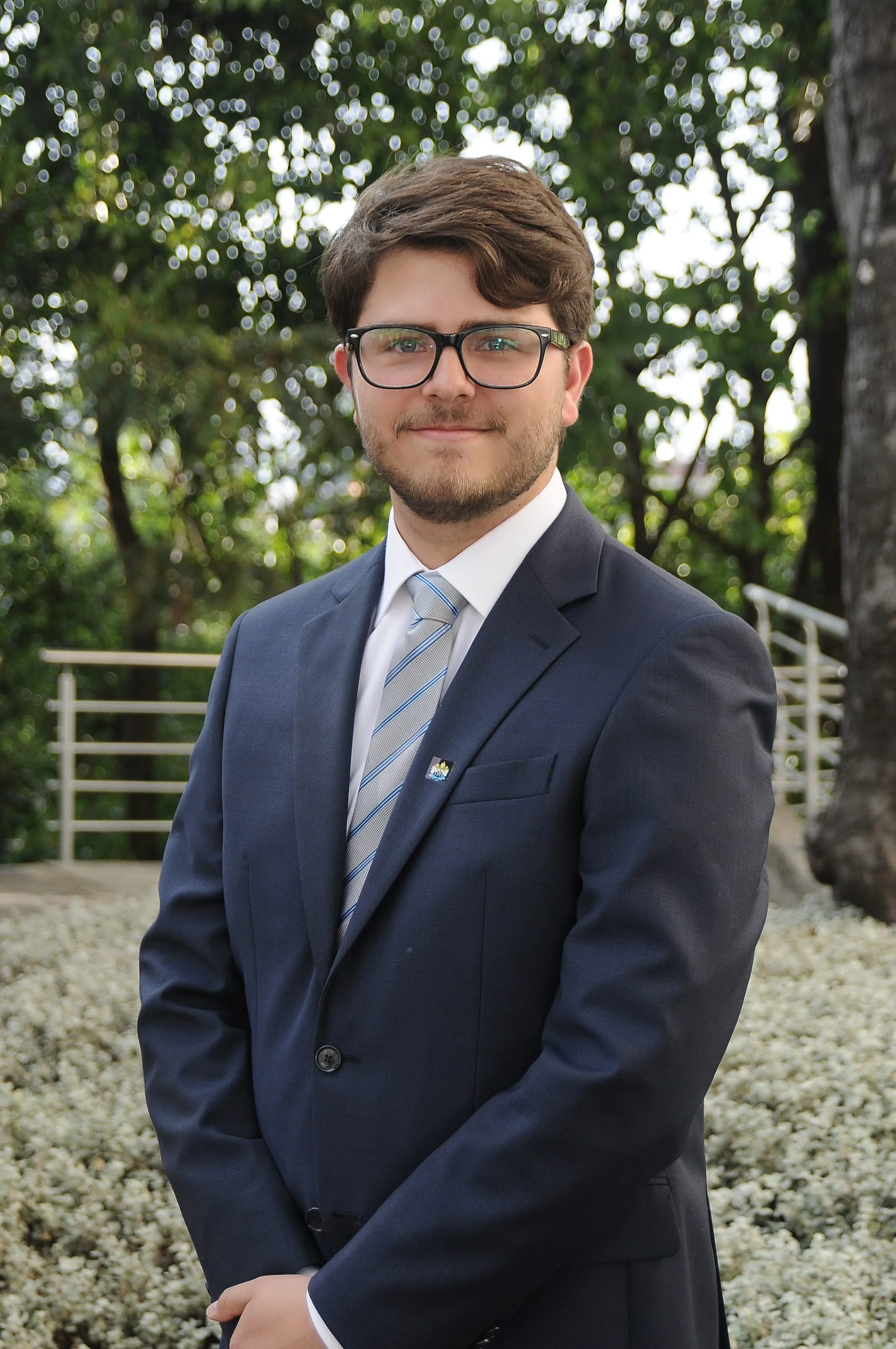 Hombre con gafas y barba, vestido con saco y corbata, posando en un entorno natural con árboles y flores.