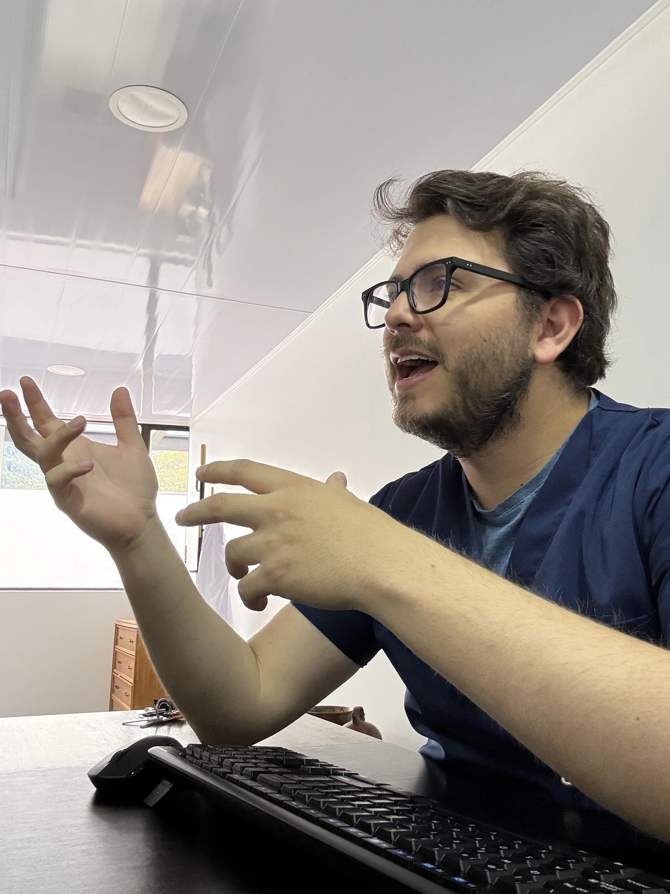 Un hombre con barba y gafas parece estar explicando algo, usando gestos con las manos, sentado frente a un teclado en una oficina o consultorio con luz natural.