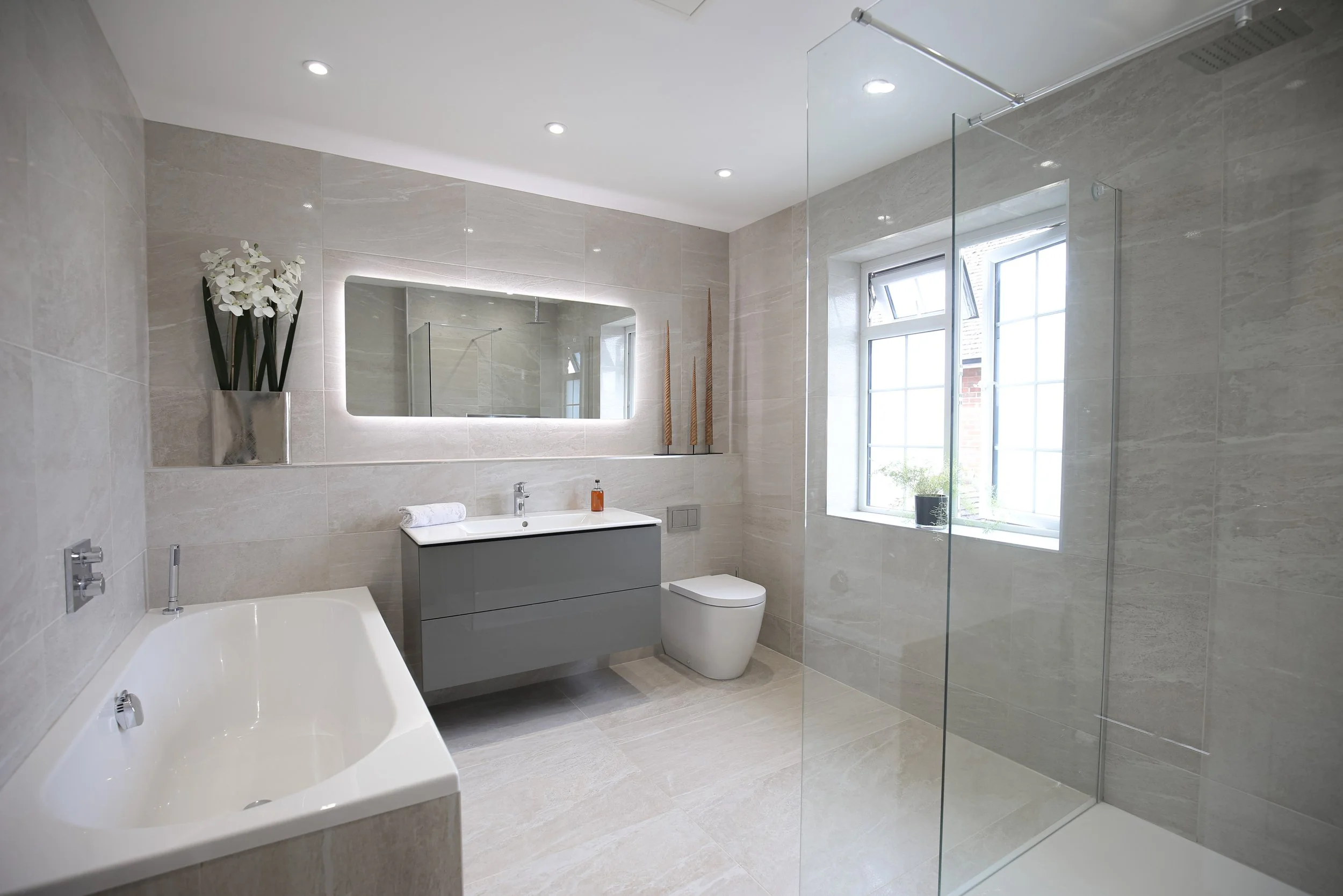 Modern bathroom with a large bathtub, floating vanity with a sink, oval mirror with backlight, toilet, window with a plant on the sill, and a shower area with glass partition.