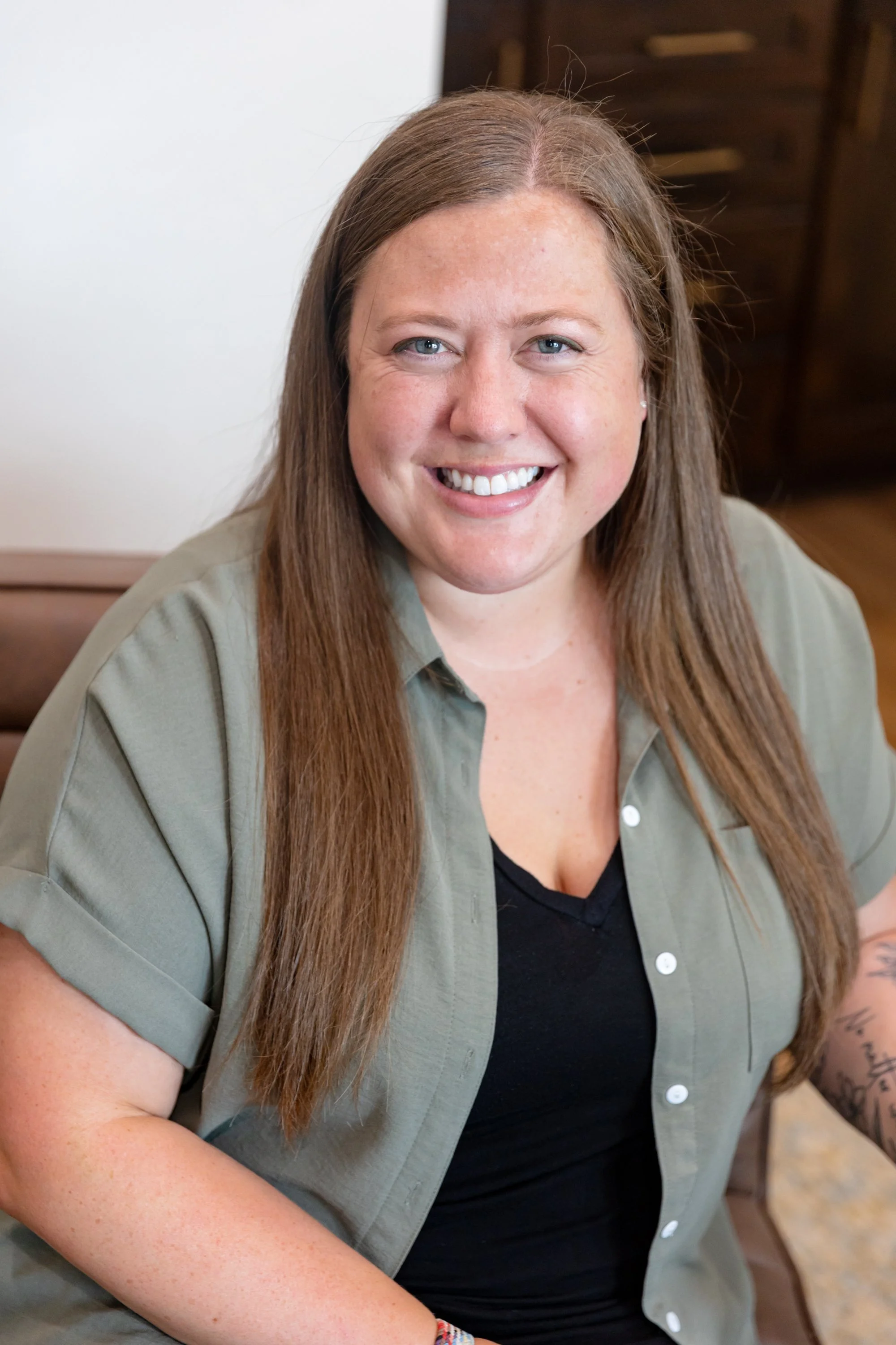 A woman with long light brown hair, wearing a black shirt and an olive green button-up shirt, smiling with white teeth, in an indoor setting with wooden furniture.