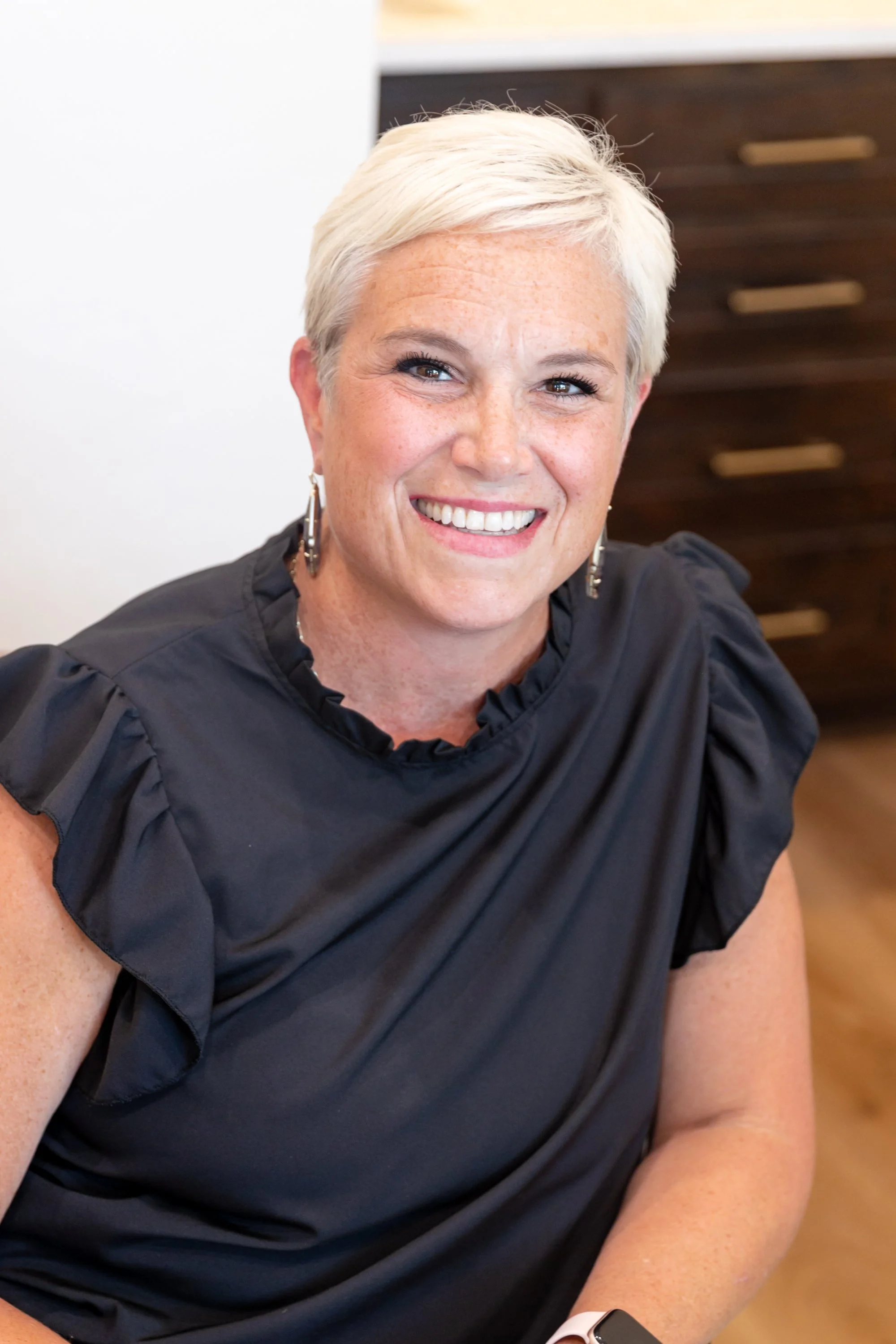Close-up of a smiling middle-aged woman with short white hair, wearing earrings and a black top with ruffled sleeves, sitting indoors.
