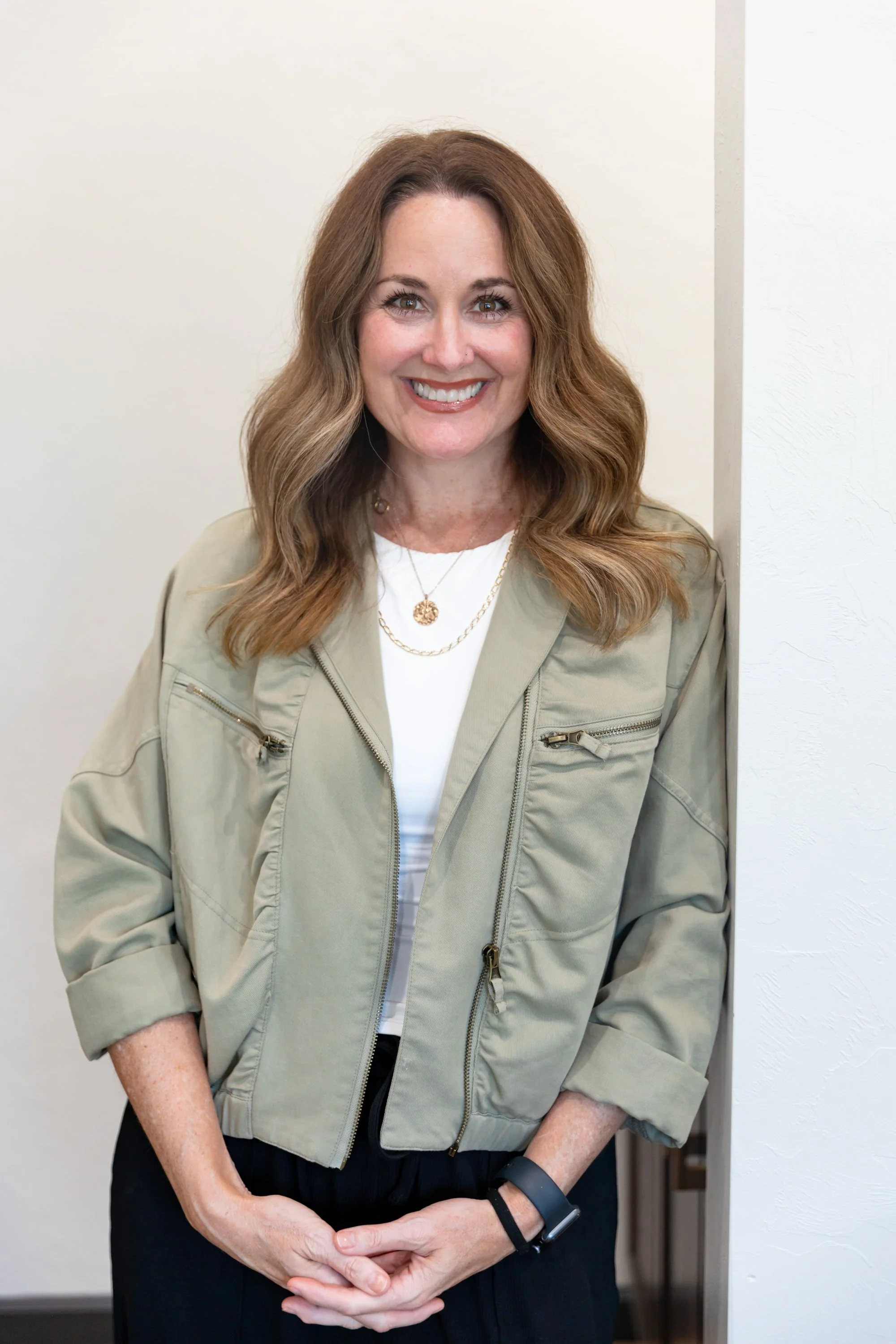 A smiling woman with long wavy brown hair, wearing a beige jacket, white shirt, black pants, a watch, and layered necklaces, standing against a plain white wall.