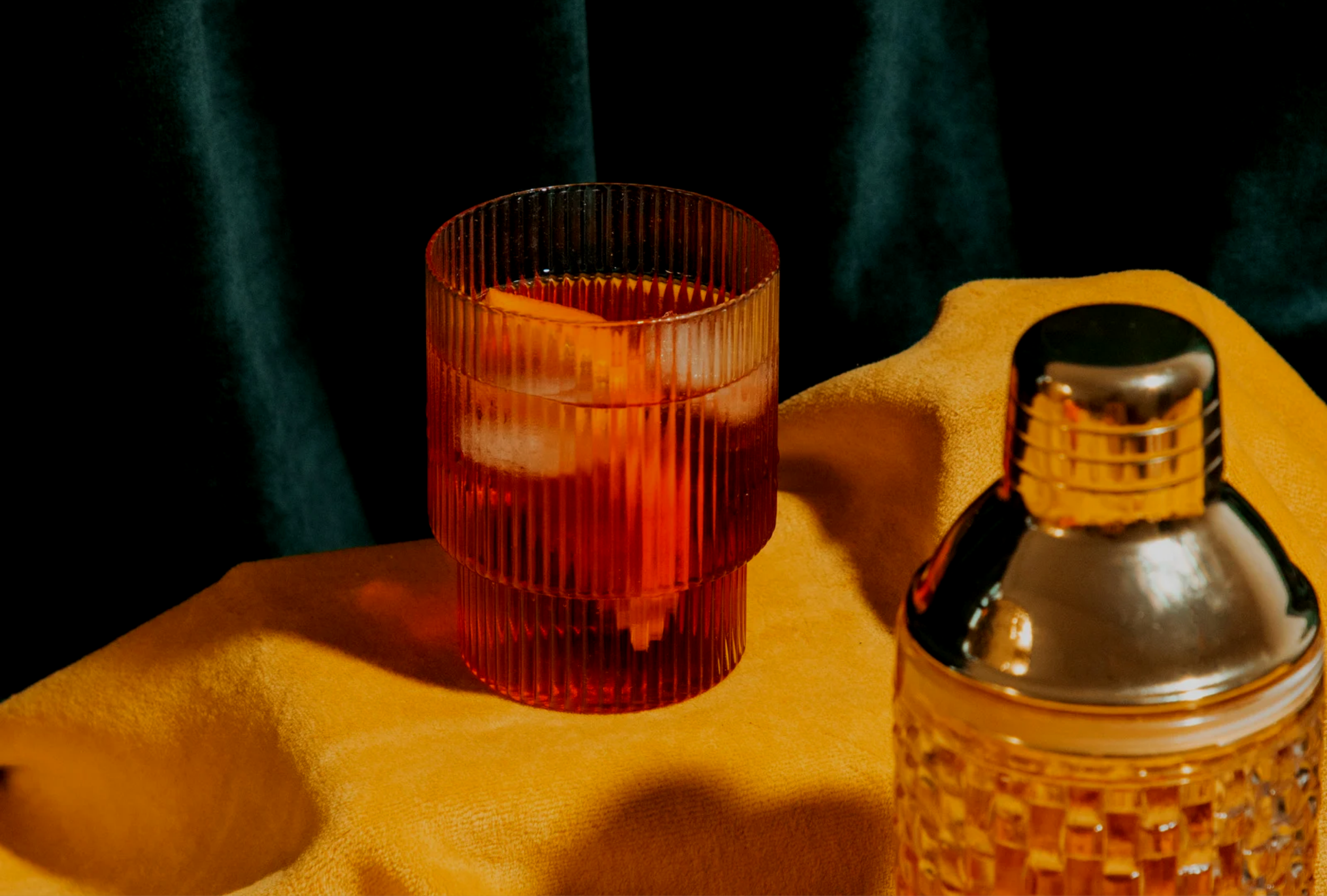 A red textured glass with ice cubes and a drink inside, placed on a yellow cloth with a dark background. A metallic spray bottle is partially visible in the foreground.