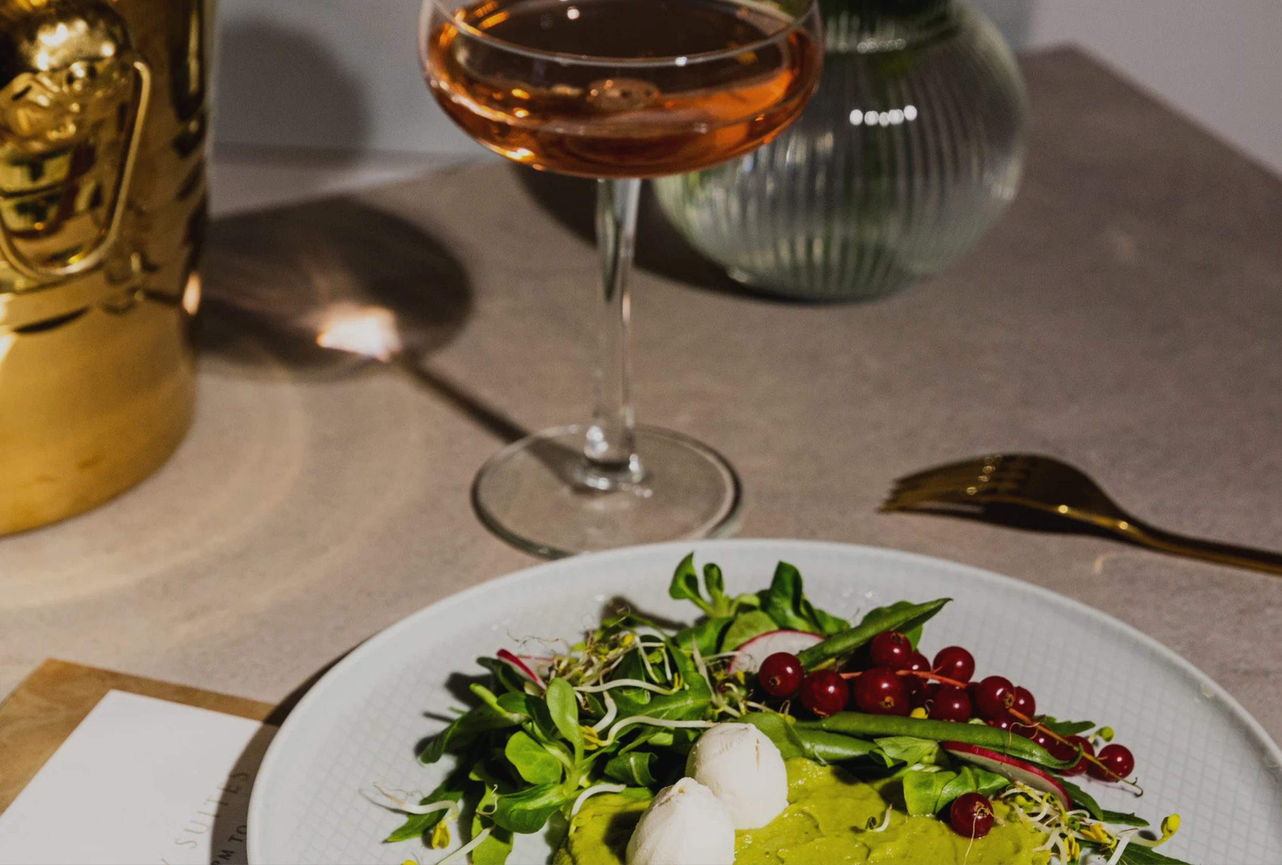 Close-up of a white plate with a salad containing greens, radishes, red currants, and dollops of avocado cream, set on a dining table with a gold fork, a glass of rosé wine, and a decorative green vase in the background.