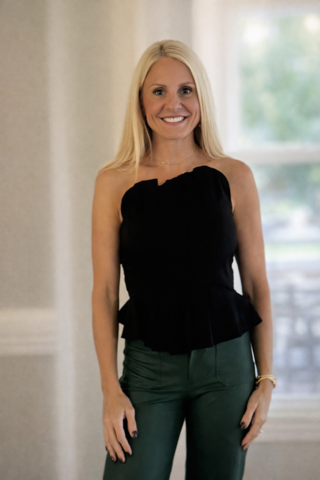 A woman with blonde hair wearing a black one-shoulder top and green pants, smiling indoors near a window.