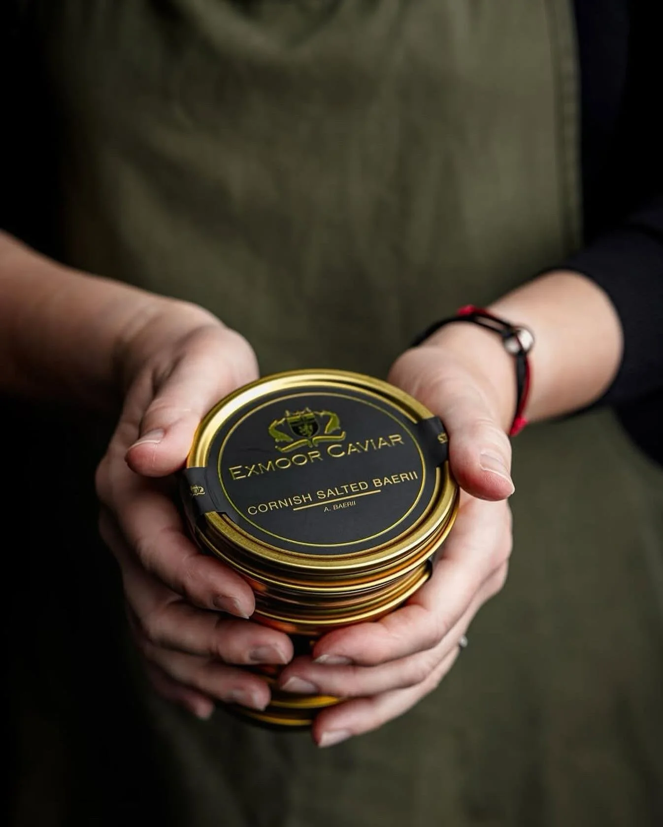 Hands holding a stack of tins of Exmoor Caviar, labeled 'Cornish Salted Baeri' with black and gold packaging.